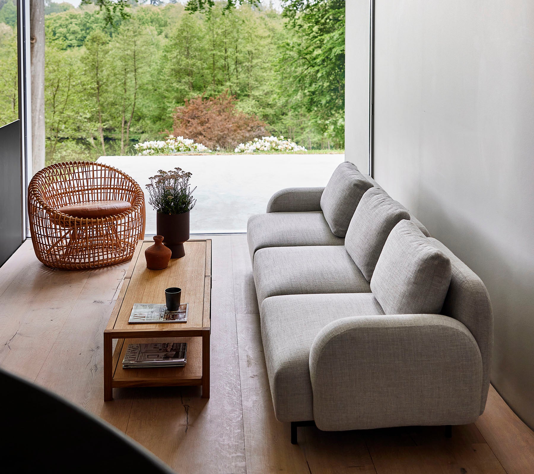 Moderne sofa i lyst stoff kombinert med et trebord og en kurvstol, omgitt av grøntområder. Naturlig sollys forbedrer den rolige atmosfæren.