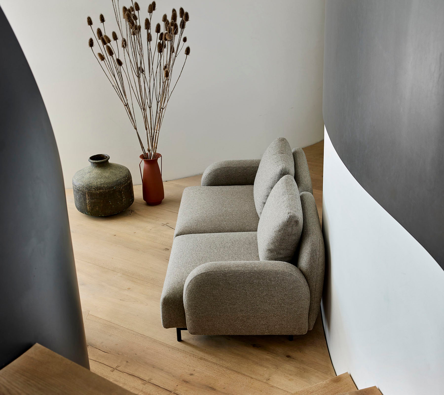 En koselig beige sofa plassert ved siden av en dekorativ vase og tørkede planter, mot en buet vegg og tregulv.