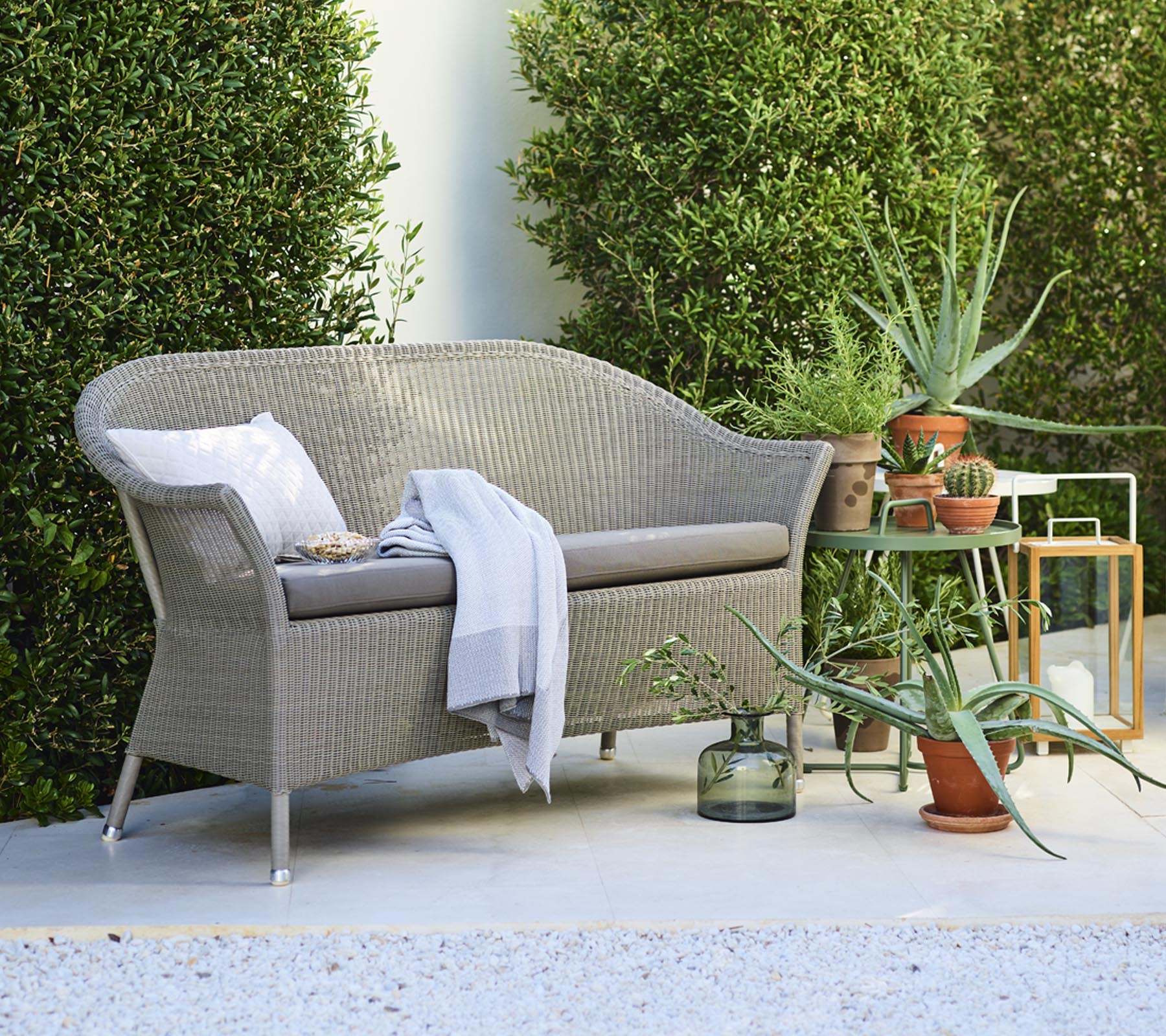 En koselig loveseat med dekorative puter, akkompagnert av potteplanter og et sidebord, omgitt av grønt.