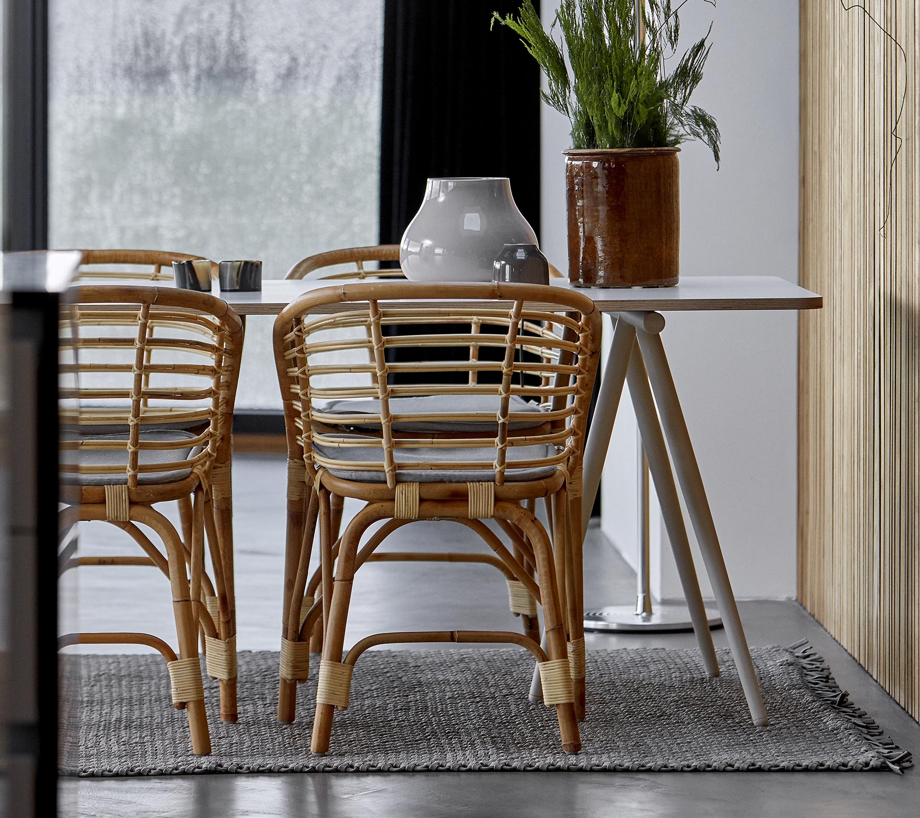 Et moderne spiseoppsett med rattanstoler, et glatt bord og dekorativt grønt i et stilrent interiør.
