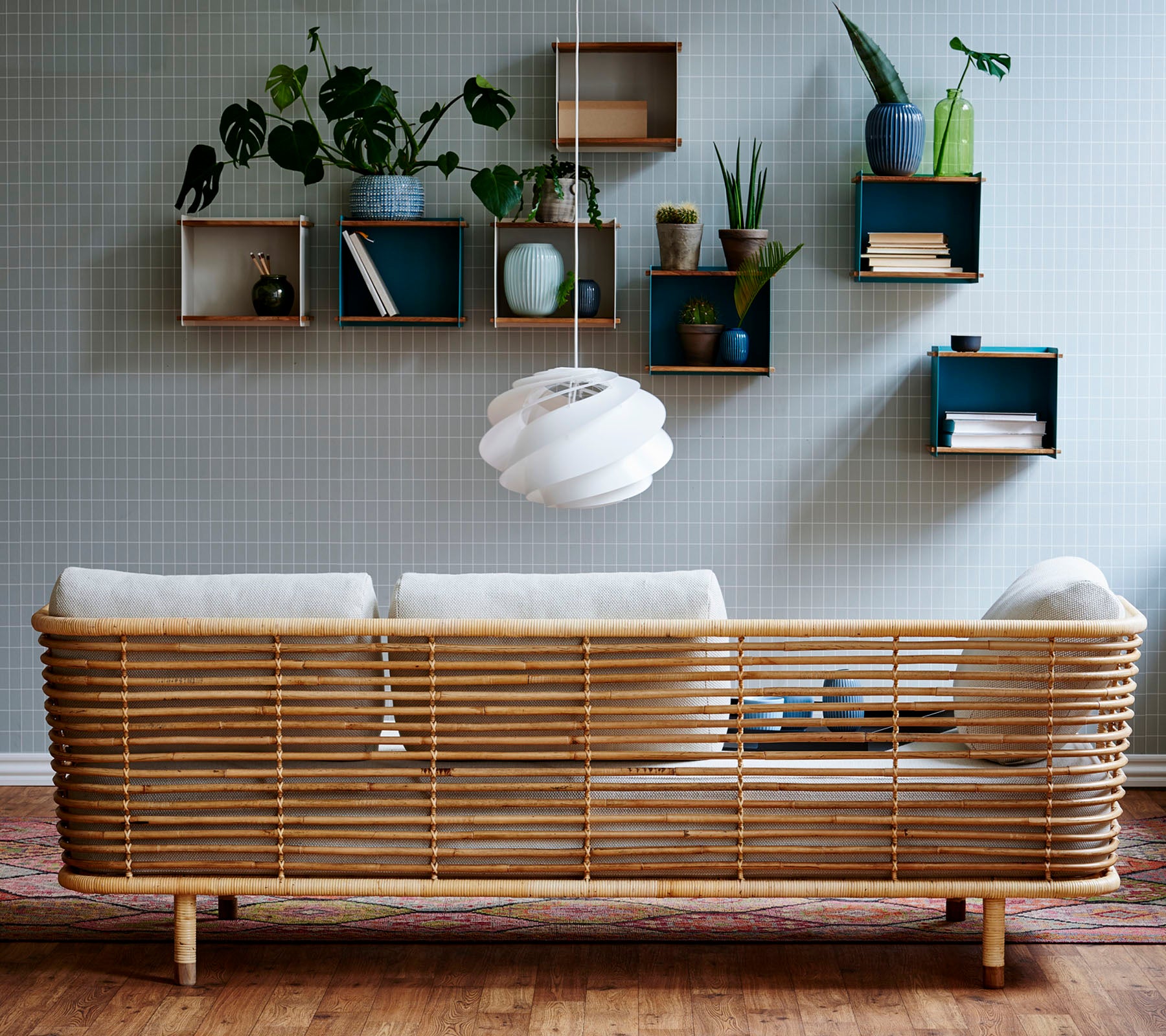 En stilfull rattan-sofa med lyse puter mot en teksturert vegg, komplementert av veggmonterte hyller som viser planter og dekorative gjenstander.