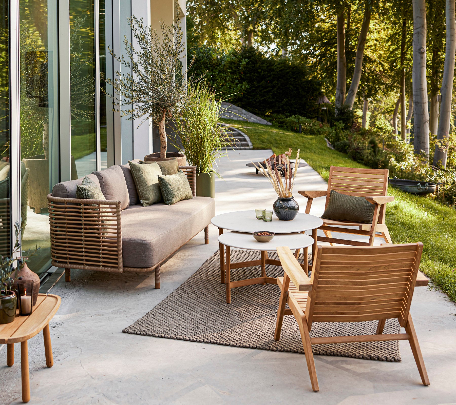Stilig uteområde med moderne sofa, trestoler, et rundt bord og dekorative planter mot en rolig bakgrunn.