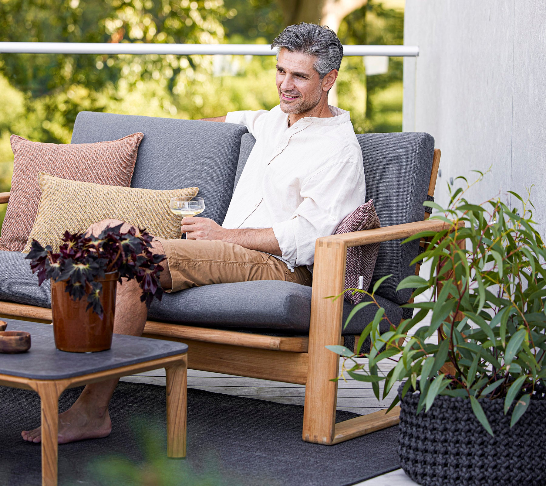 En mann nyter en drink mens han sitter på en komfortabel sofa, omgitt av planter og dekorative puter.