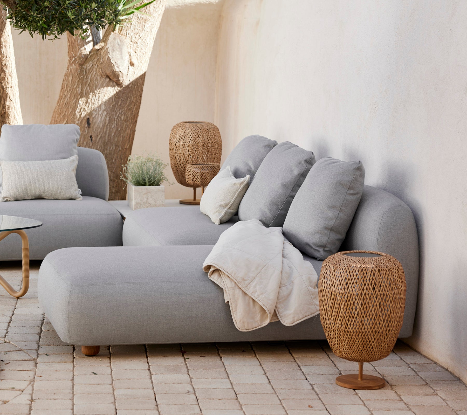 Moderne grå sofa med puter og et vevd sidebord i en minimalistisk stueløsning.