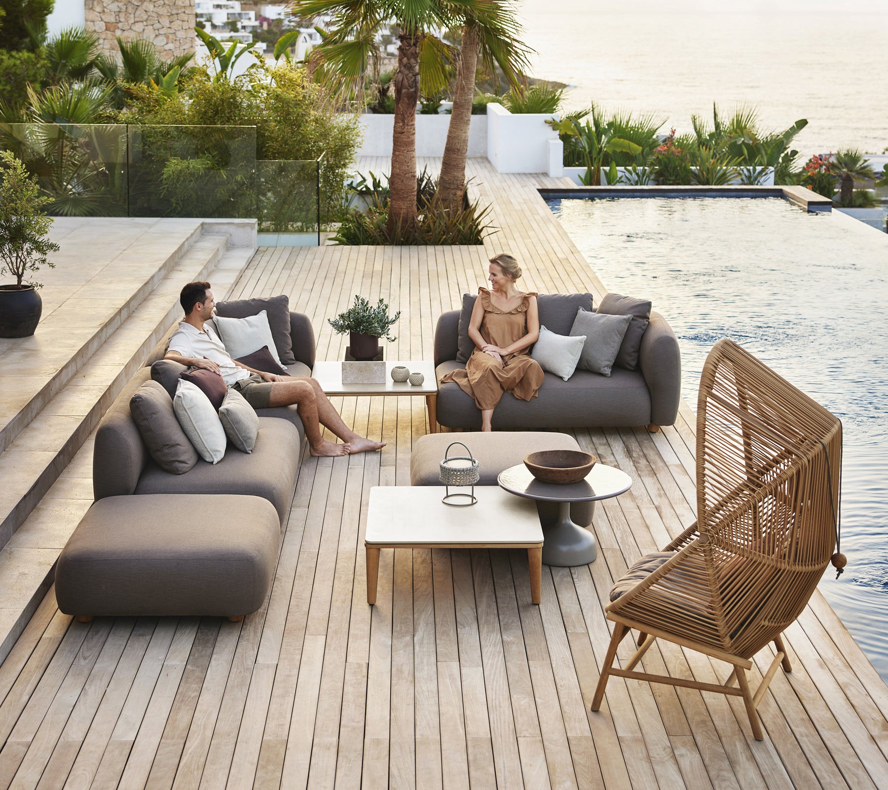 Moderne utendørs loungeområde med grå sofaer, et hvitt bord og en rattanstol.