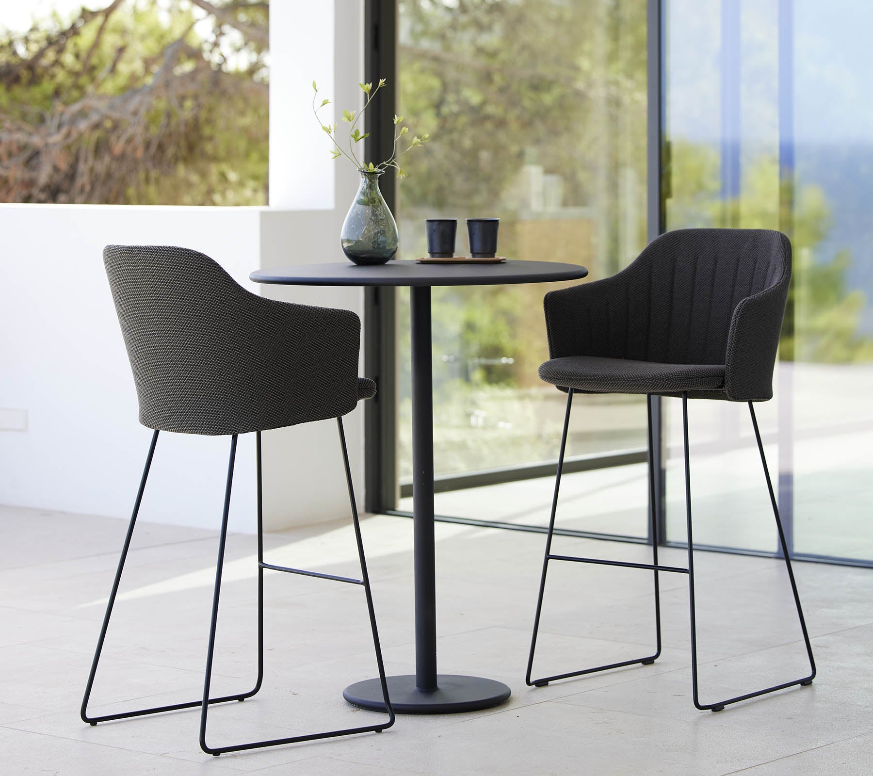 To stilige stoler parret med et elegant barbord, komplementert med en vase og kopper, som skaper en moderne og innbydende sitteordning.