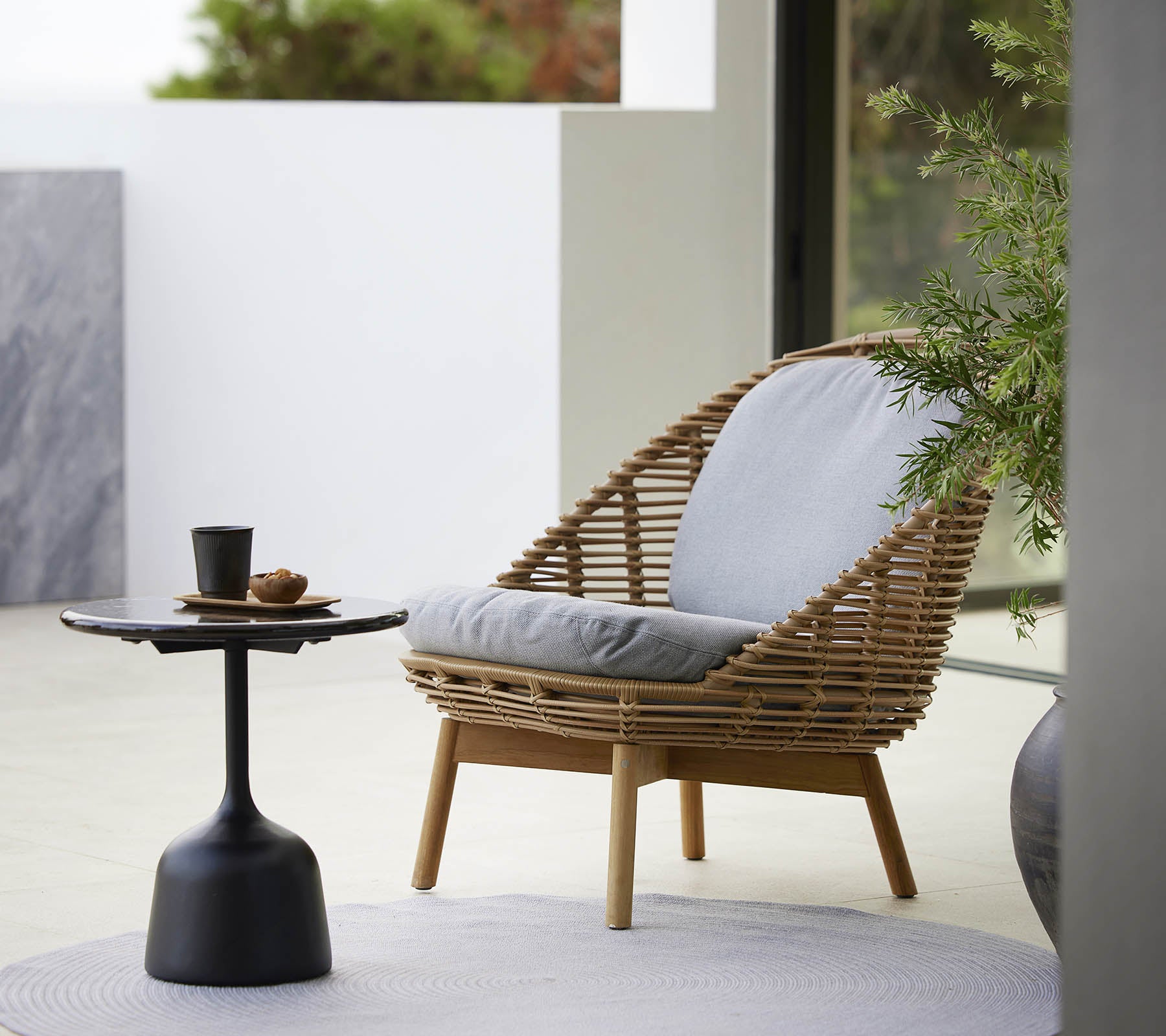 En stilfull lenestol med vevd design og myke puter, akkompagnert av et slankt sidebord og en liten plante. Ideell for avslapning.