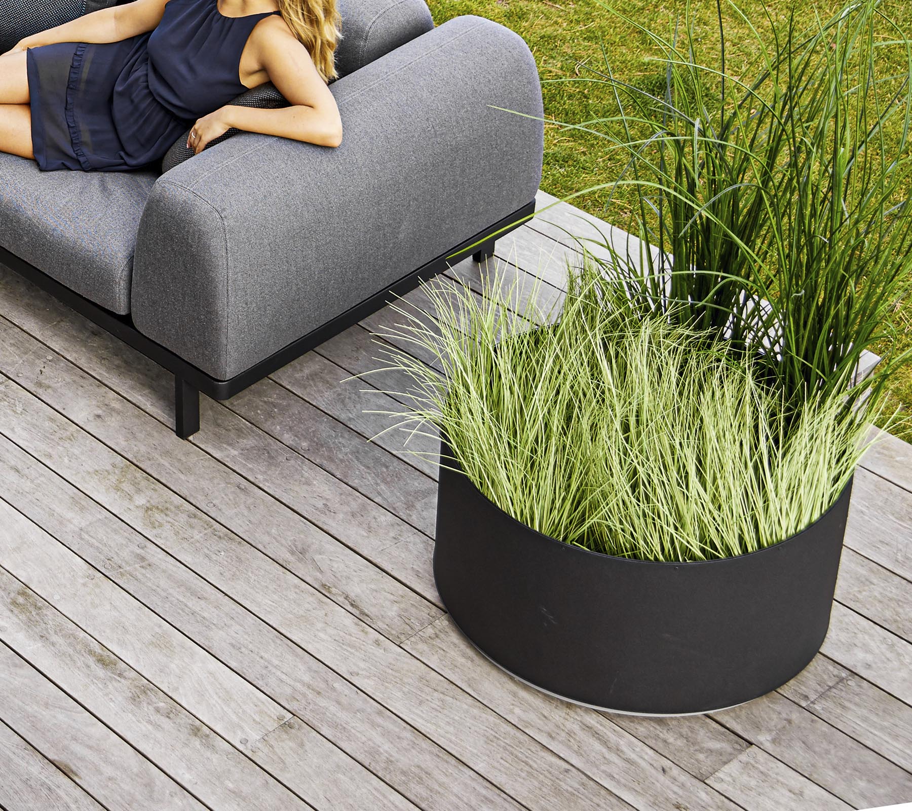 En stilfull grå sofa ved siden av en sirkulær svart blomsterpotte fylt med høyt grønt gress, plassert på treflo