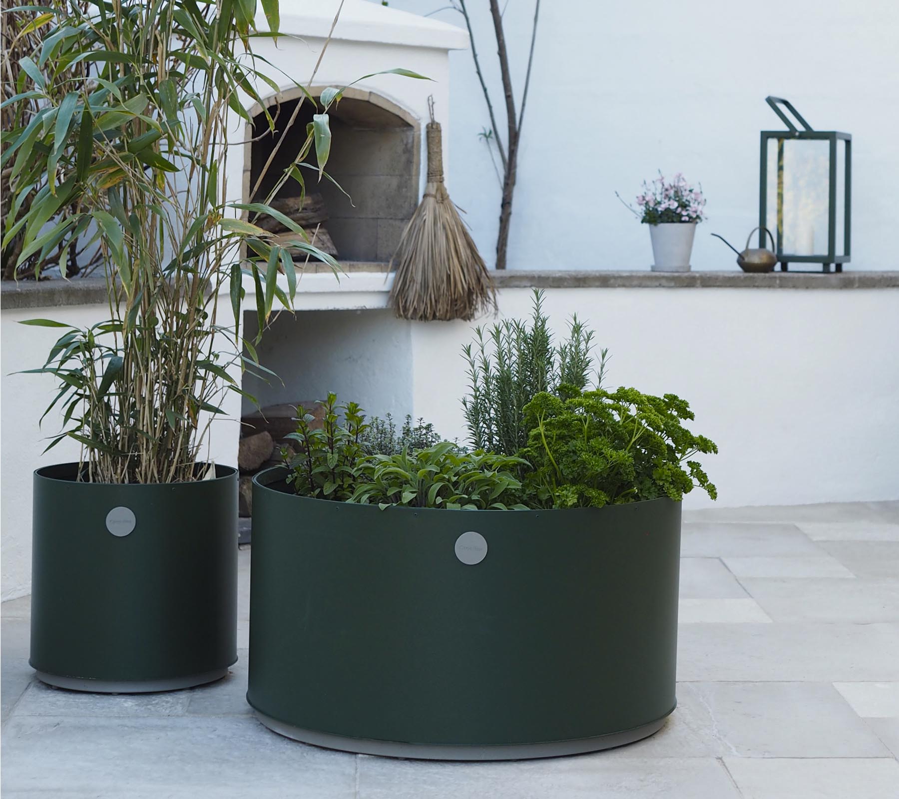 To runde plantekasser i mørkegrønn, den ene med høye planter og den andre fylt med forskjellige urter, står på en lys steinoverflate.