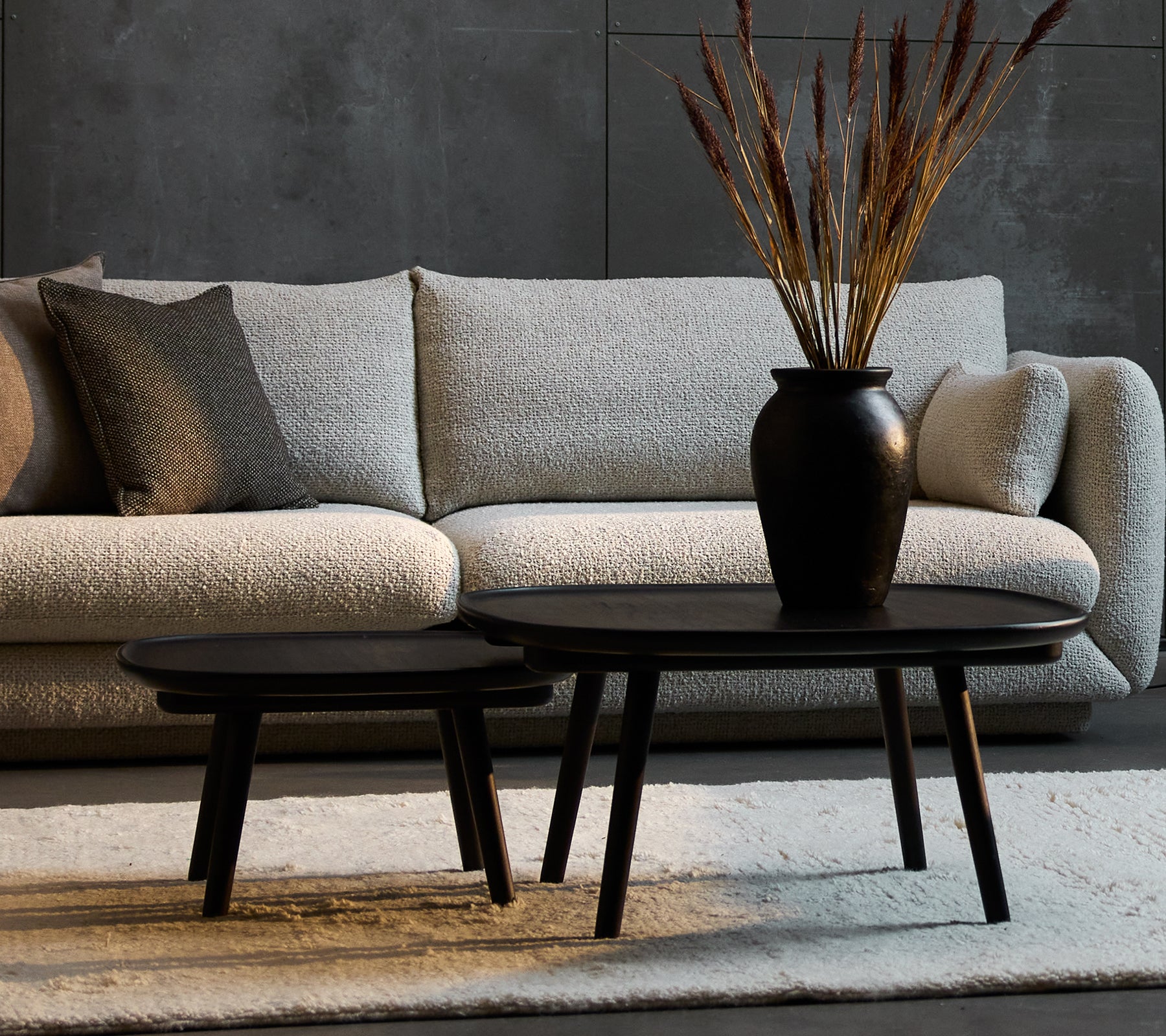 To elegante sorte sofabord i ulike størrelser, komplementert med en dekorativ vase med tørkede planter, plassert mot en koselig sofa.