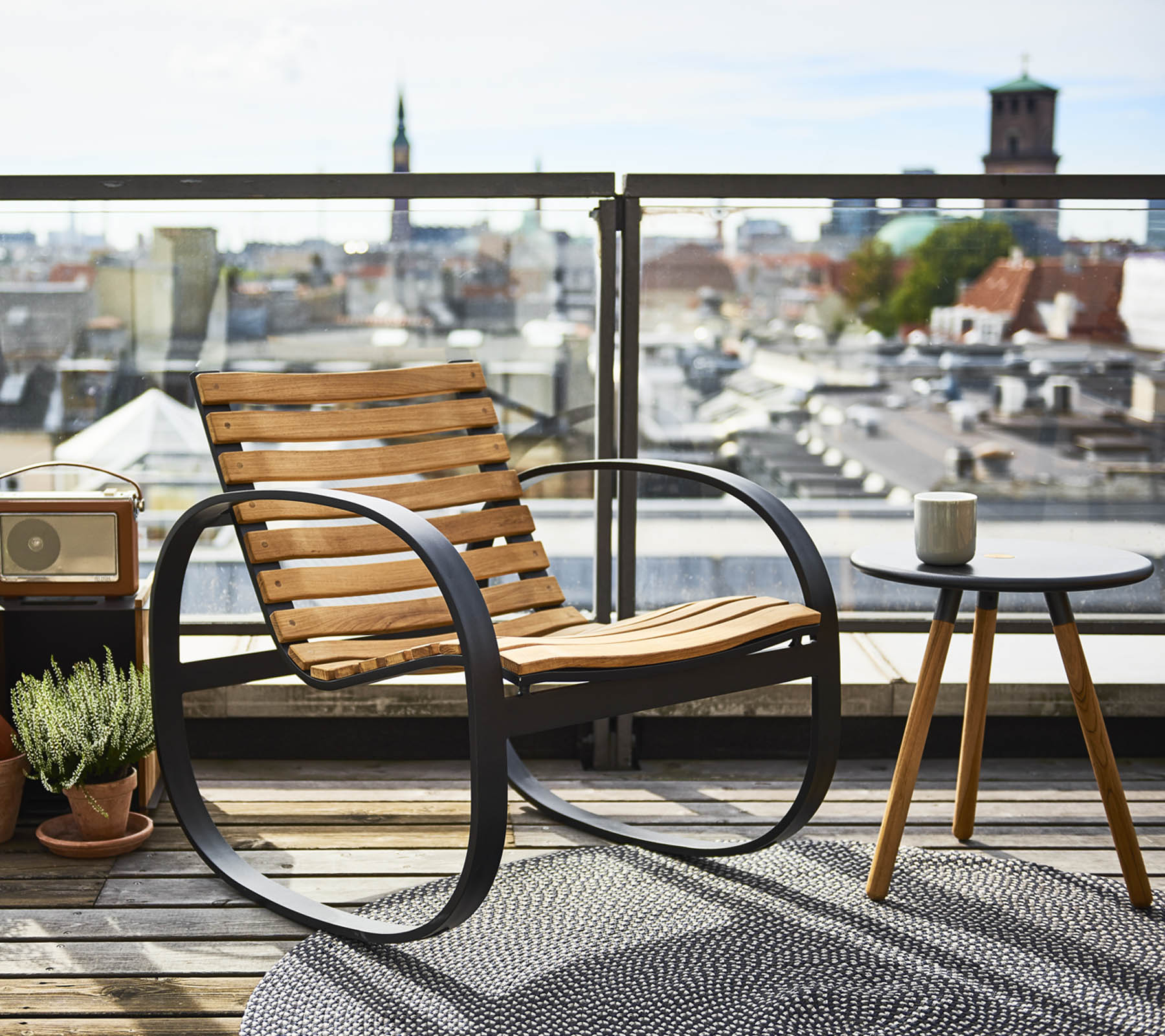 En stilig gyngestol ved siden av et lite sidebord, mot en panoramautsikt over byen, pyntet med en plante og et teppe.