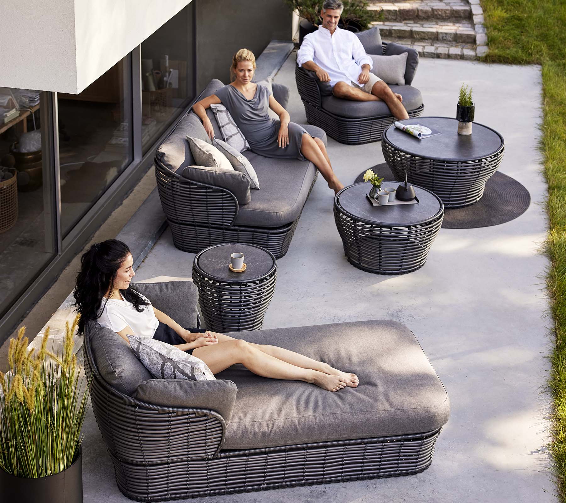 Moderne loungeområde med en kurv-dagbed, en 3-seters sofa og loungestoler, supplert med runde bord og dekorative planter.