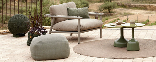 Stilig sitteområde med en komfortabel stol, rundt bord, pouf og dekorative tilbehør arrangert på et strukturert teppe.