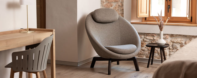 Koselig sitteområde med en moderne stol med avrundet design, et lite sidebord og et minimalistisk skrivebord. Naturlig lys strømmer inn gjennom et vindu.