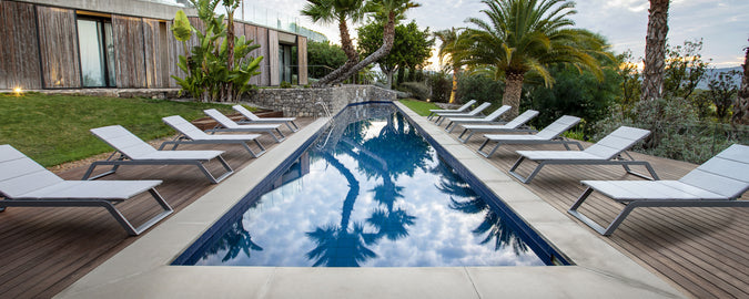 En rolig bassengscene med en rad av hvite solsenger ved siden av et stille vann, omkranset av frodig vegetasjon og palmer.