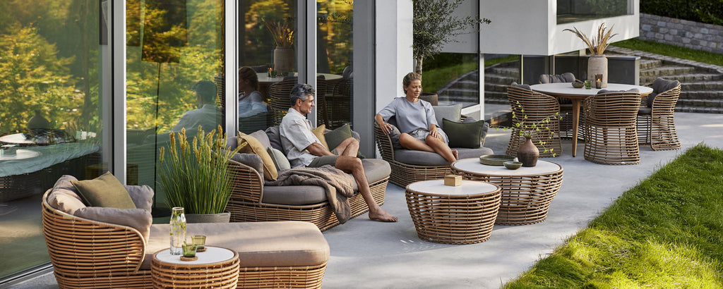 Moderne rattanstoler med grå puter og et kaffebord i en moderne utendørs loungeinnstilling.