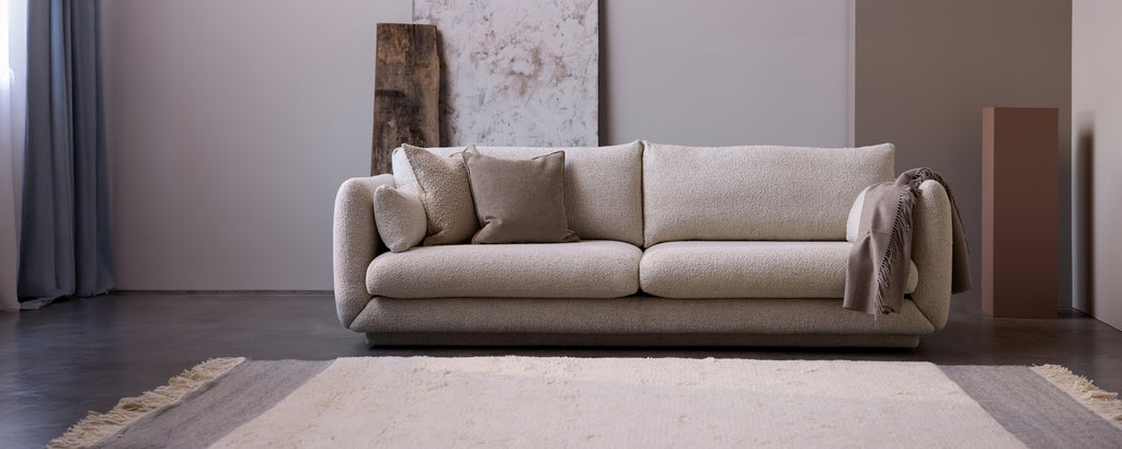 Moderne beige sofa med puter og et grått pledd i en minimalistisk stueinnredning.