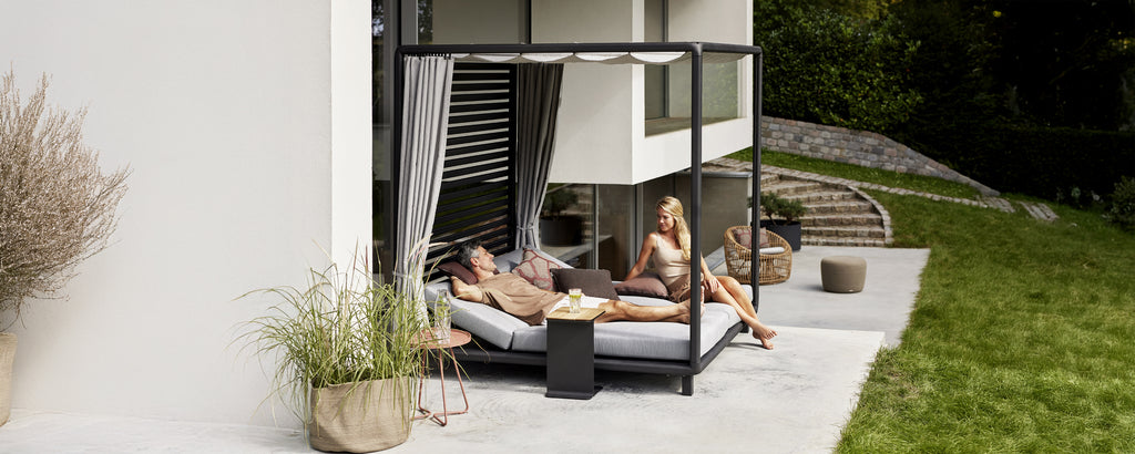 Moderne ute loungeområde med en svart daybed med grå puter og hvite gardiner.
