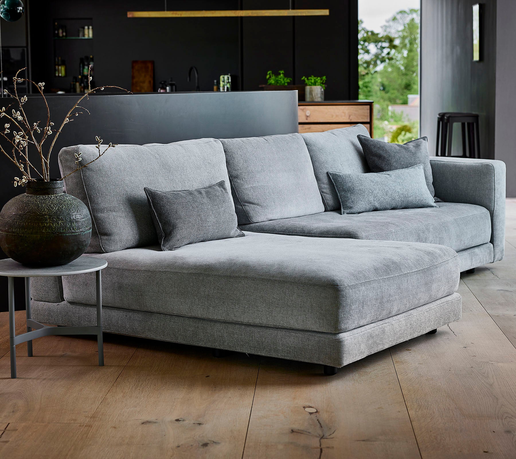Grå seksjon sofa med teksturert stoff og dekorative puter, sammen med et rundt sidebord og en stilfull vase.