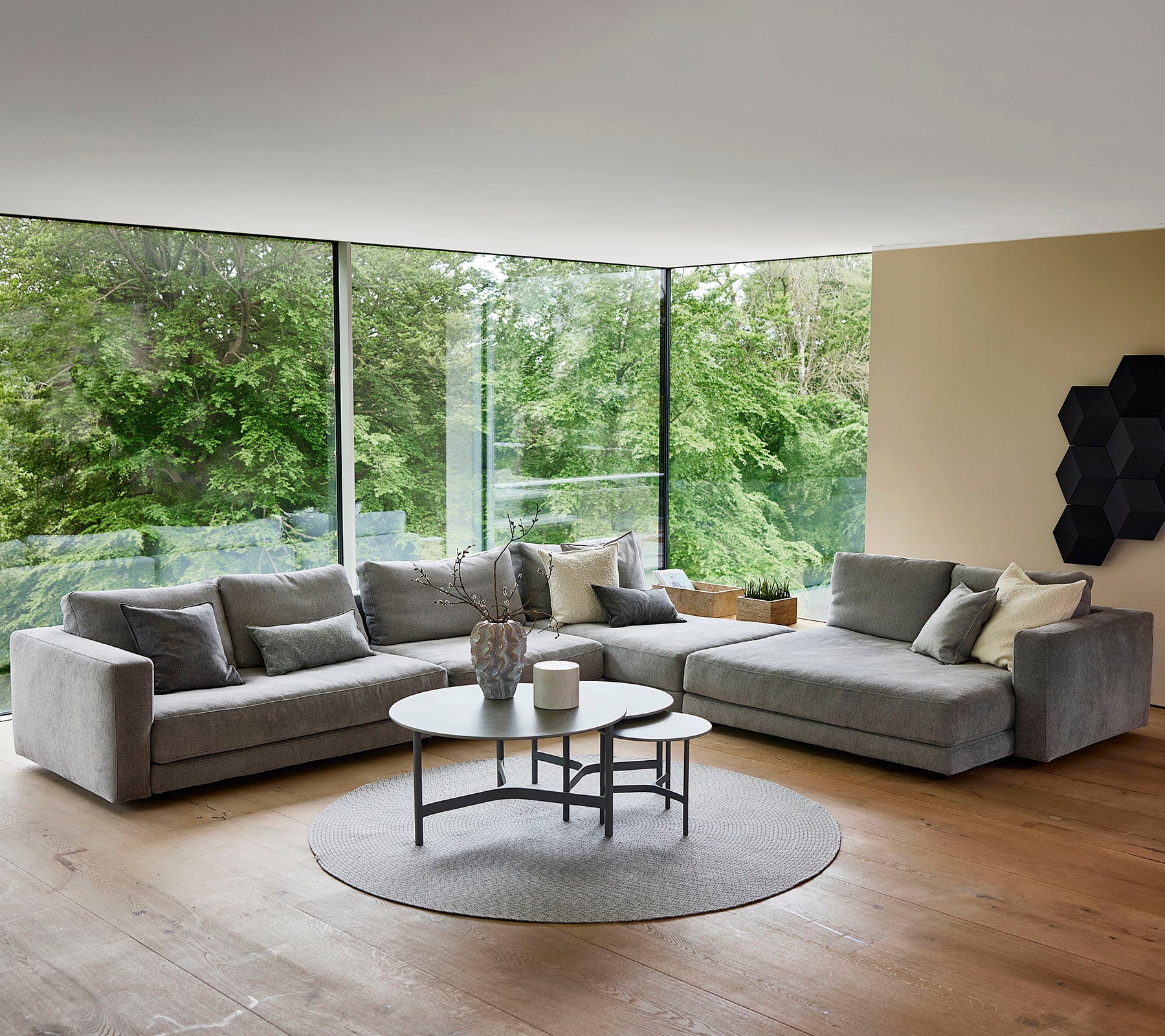 A stylish, modern living space featuring a plush gray sectional sofa, a round coffee table, and large windows showcasing greenery outside.
