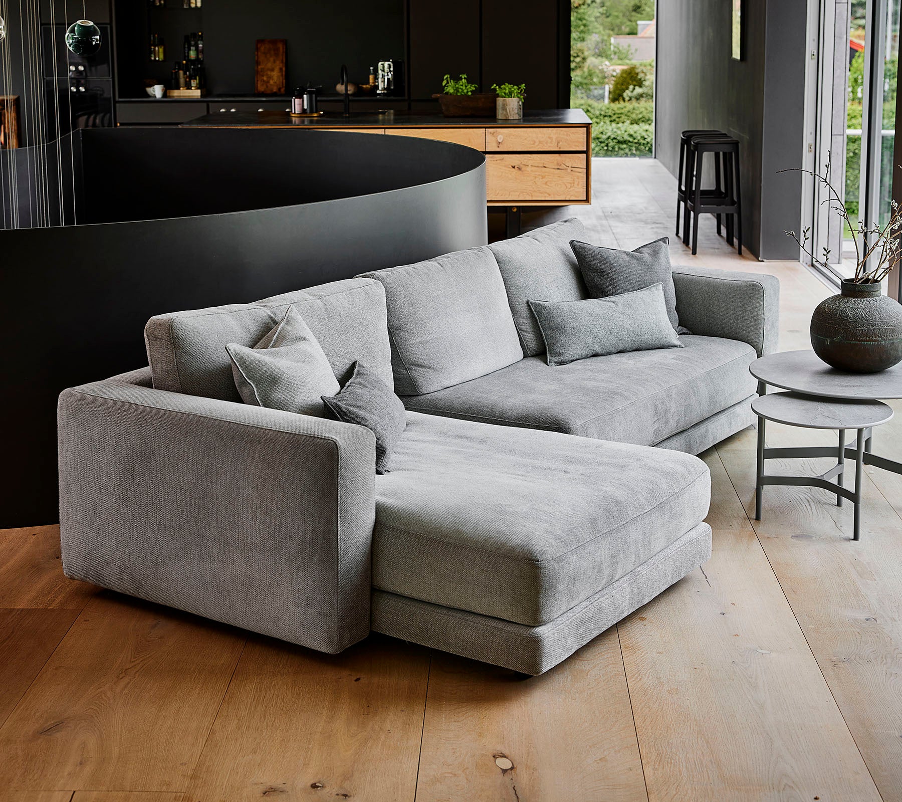 Moderne grå seksjonsofa med flere puter, akkompagneres av et rundt salongbord og en dekorativ vase. Enkelt, elegant design.