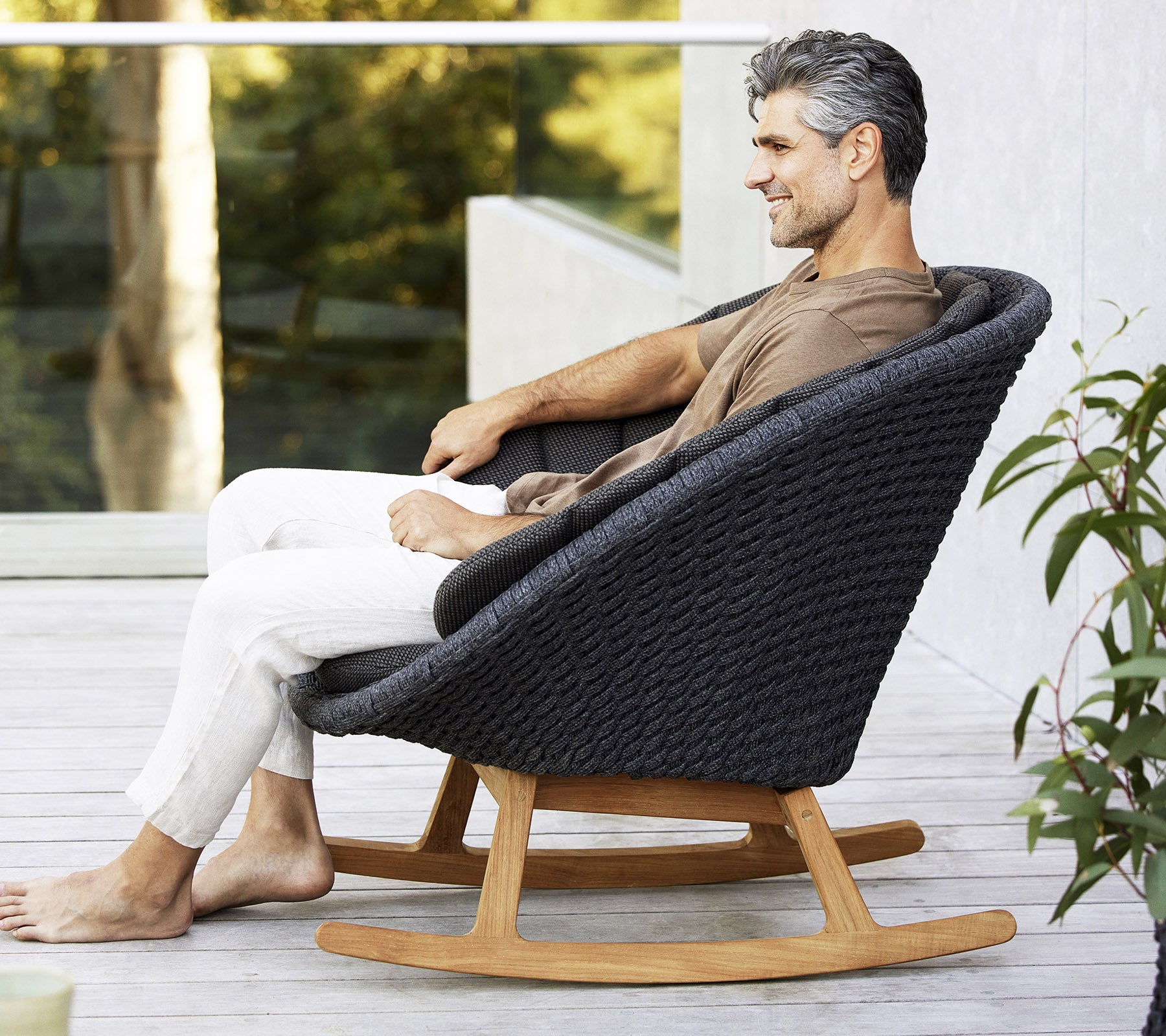 En mann sitter avslappet i en stilfull, moderne gyngestol med et vevd design og en base av naturlig tre, omringet av grønt.