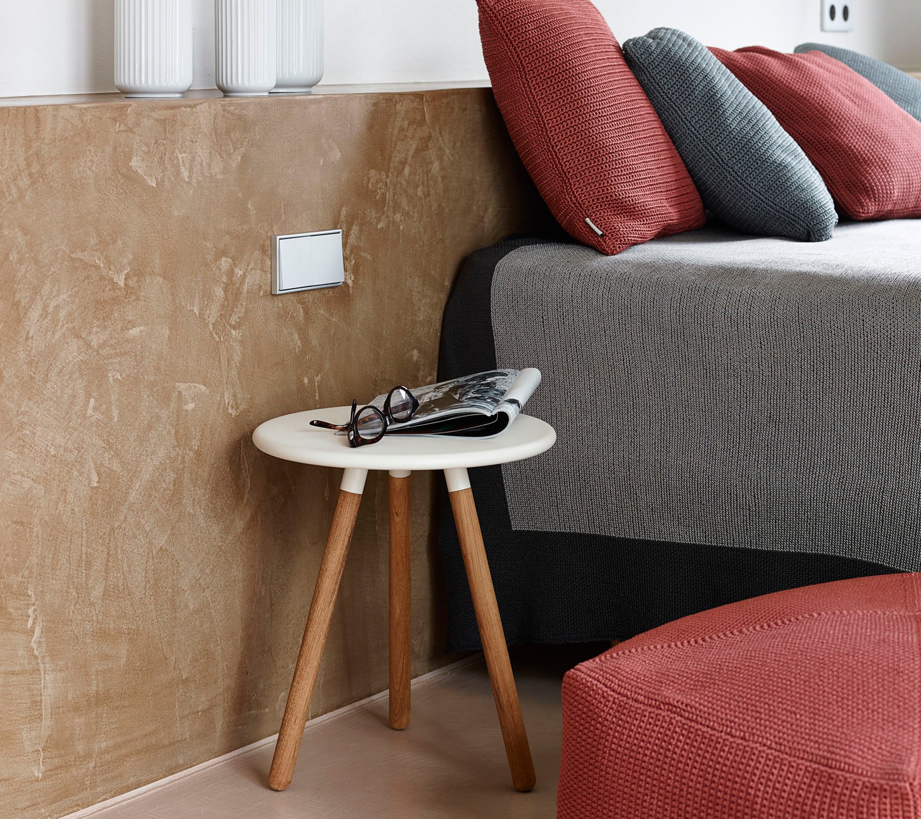 En koselig soveromscene med et lavt sidebord med briller og et magasin ved siden av en pent redd seng med teksturerte puter.