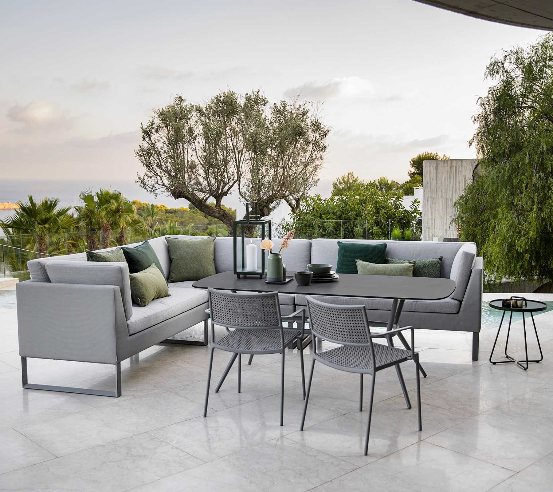 Moderne utendørs sitteområde med en stilig seksjonssofa, et spisebord og metallstoler omkranset av grøntområder.