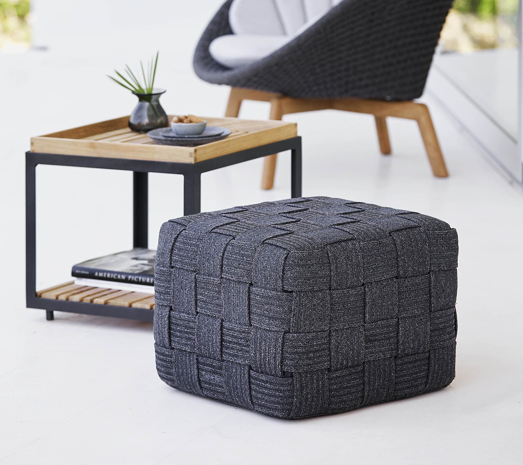 En teksturert grå vevd pouf plassert nær et minimalistisk sidebord i tre, med små dekorative gjenstander.