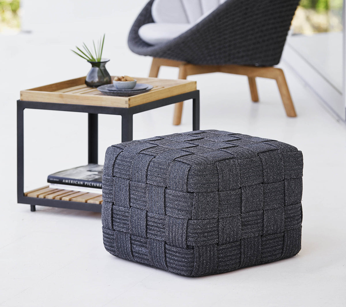 En teksturert grå vevd pouf plassert nær et minimalistisk sidebord i tre, med små dekorative gjenstander.