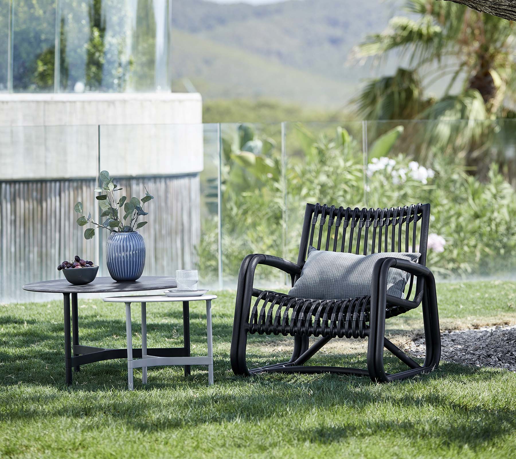 Moderne lenestol med et mønstrete design, ledsaget av et sidebord med en vase og en potteplante, plassert i et naturlandskap.