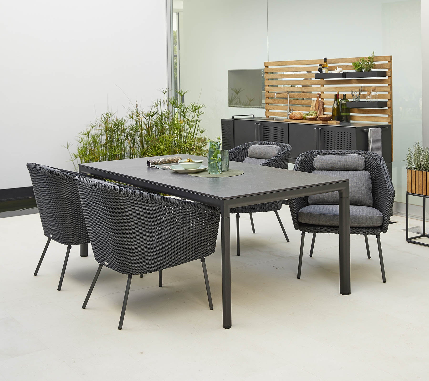 Moderne utendørs spisebord med et stilrent bord og fire stilige stoler med komfortable puter, omgitt av grønne planter.