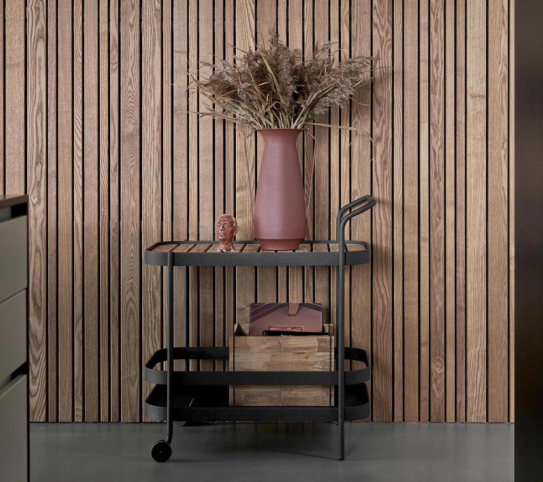 En stilren vogn med en mørk finish, som har trelagrede oppbevaringsbokser i tre og en rosa vase med tørkede blomster.