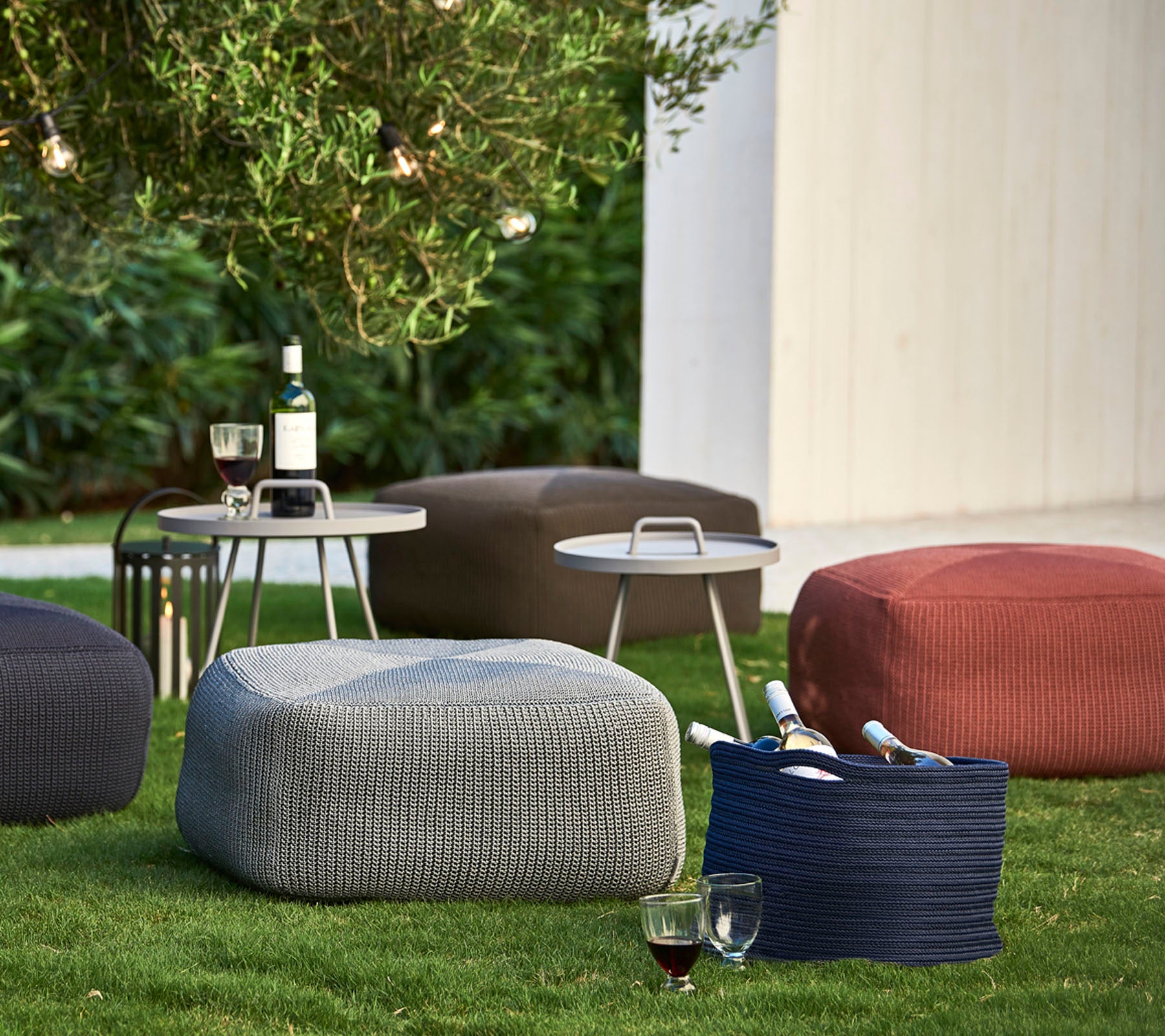 Teksturerte poufs i ulike farger plassert på gresset, ledsaget av et lite bord med drikke og vinflasker.