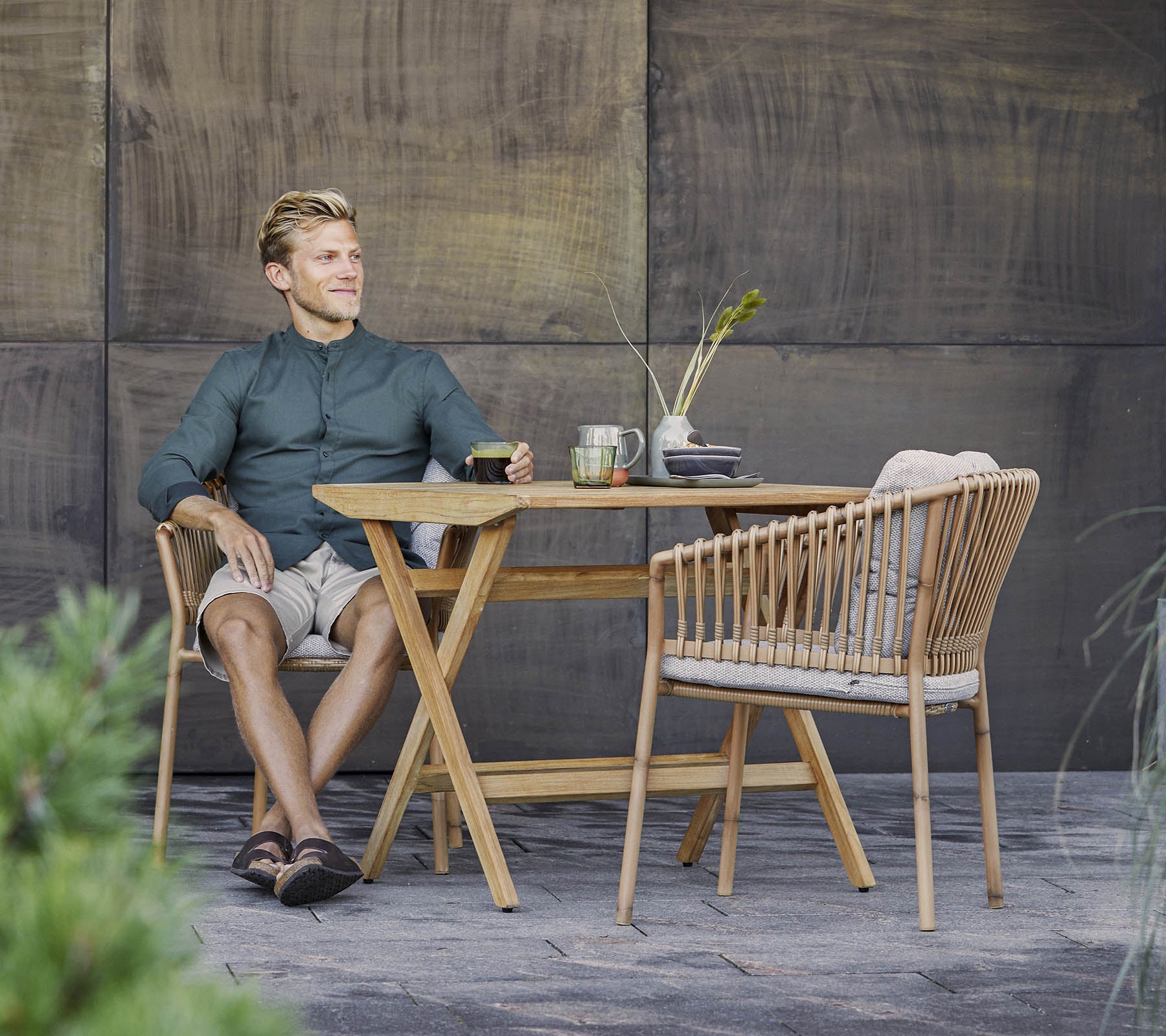 En mann sitter avslappet ved et stilfullt tremøbel med en lett drink, omringet av grønt og en teksturert bakgrunn.