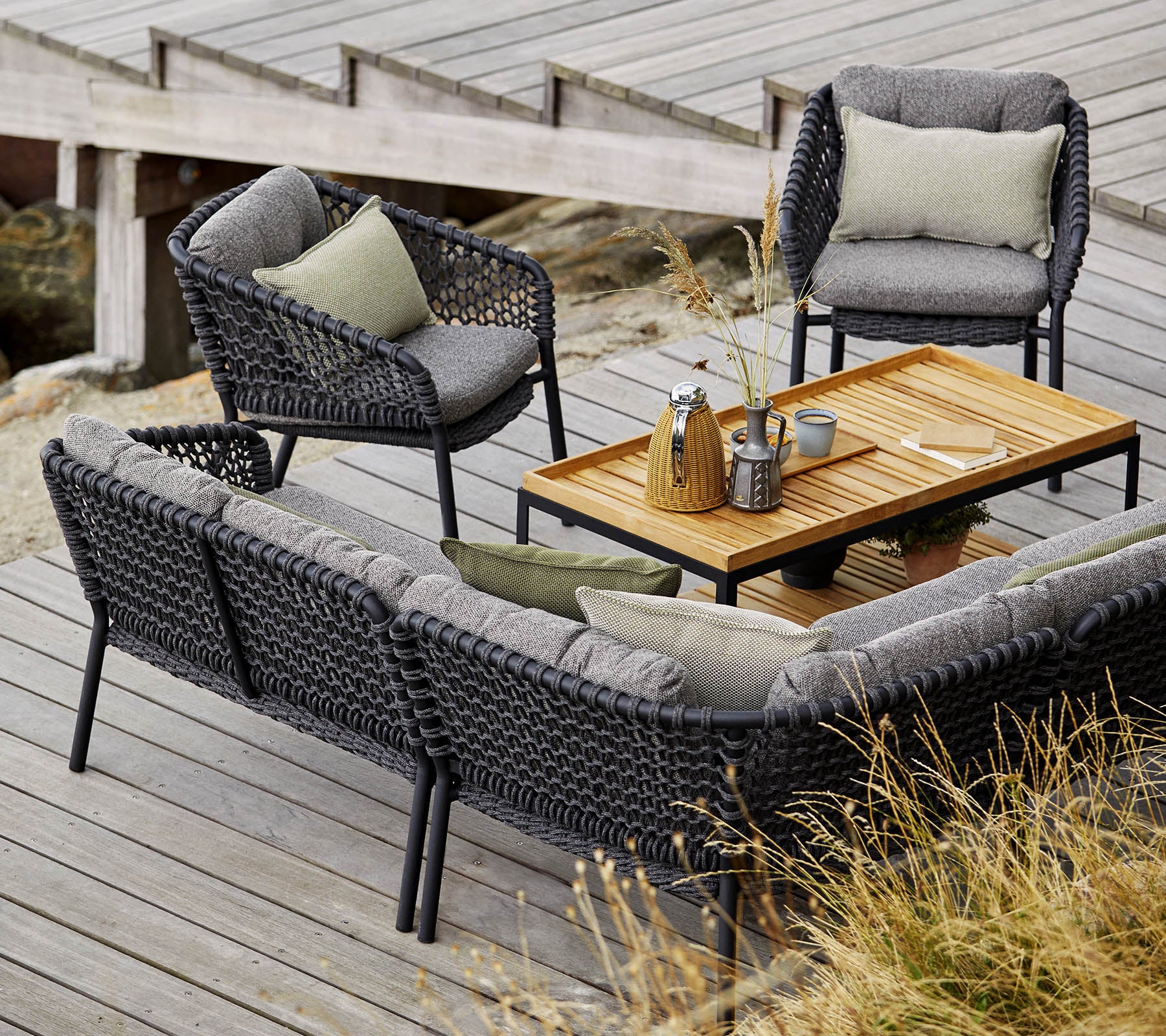 Stilsett for utendørs sitteplasser med en koselig sofa, to stoler og et trebord, accentuert med myke puter og dekorative gjenstander.