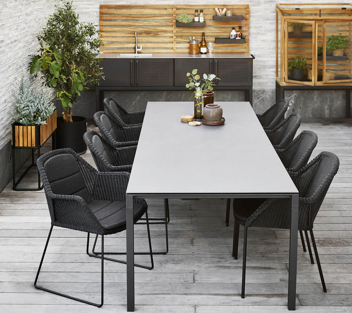 Et moderne middagsoppsett med et langt bord omgitt av slanke svarte stoler, komplementert av dekorative planter og en trebakgrunn.