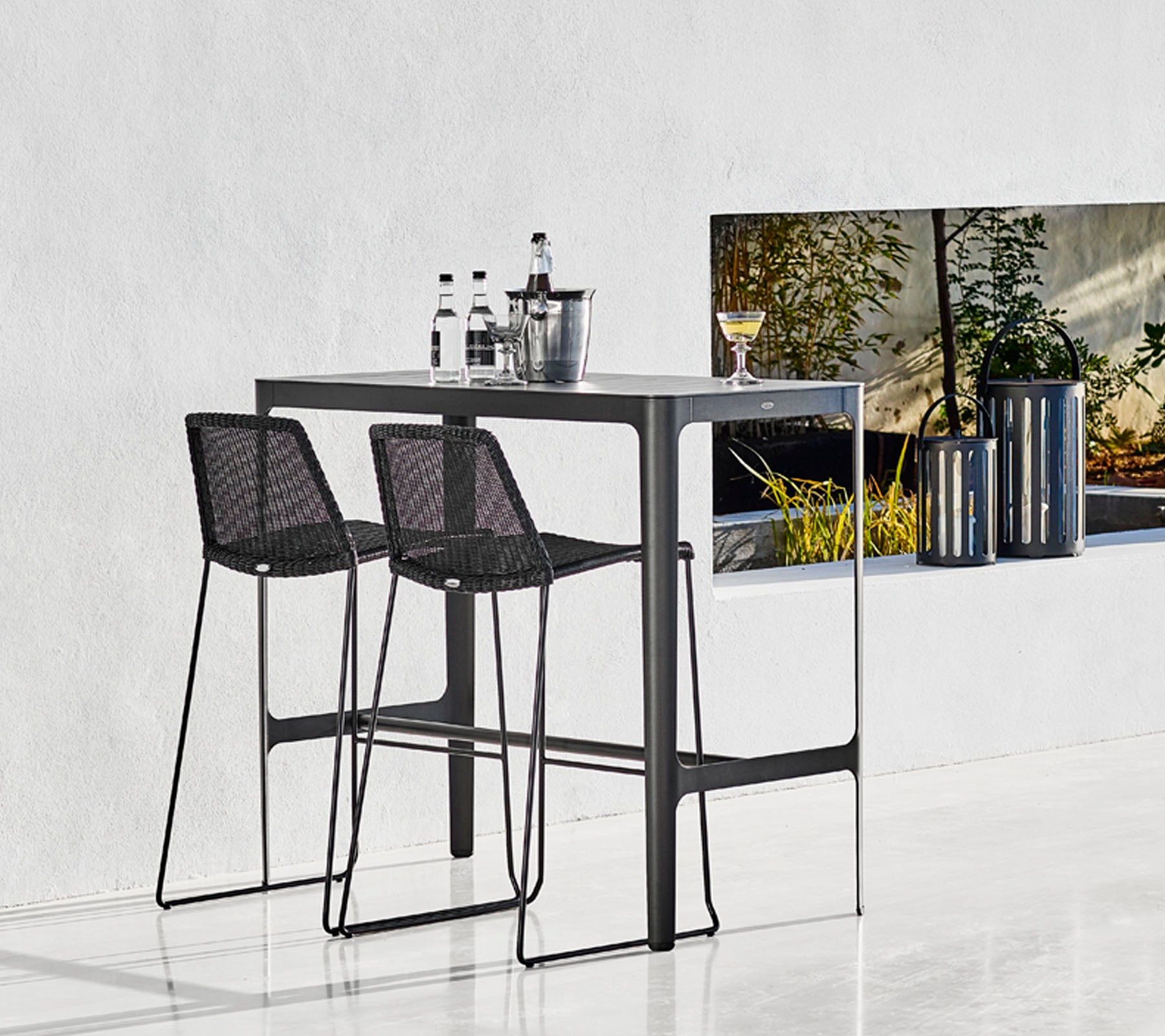 Elegant barbord med to svarte vevde barstoler, akkompagnert av glassaker og en stilfull bøtte, satt mot en minimalistisk bakgrunn.