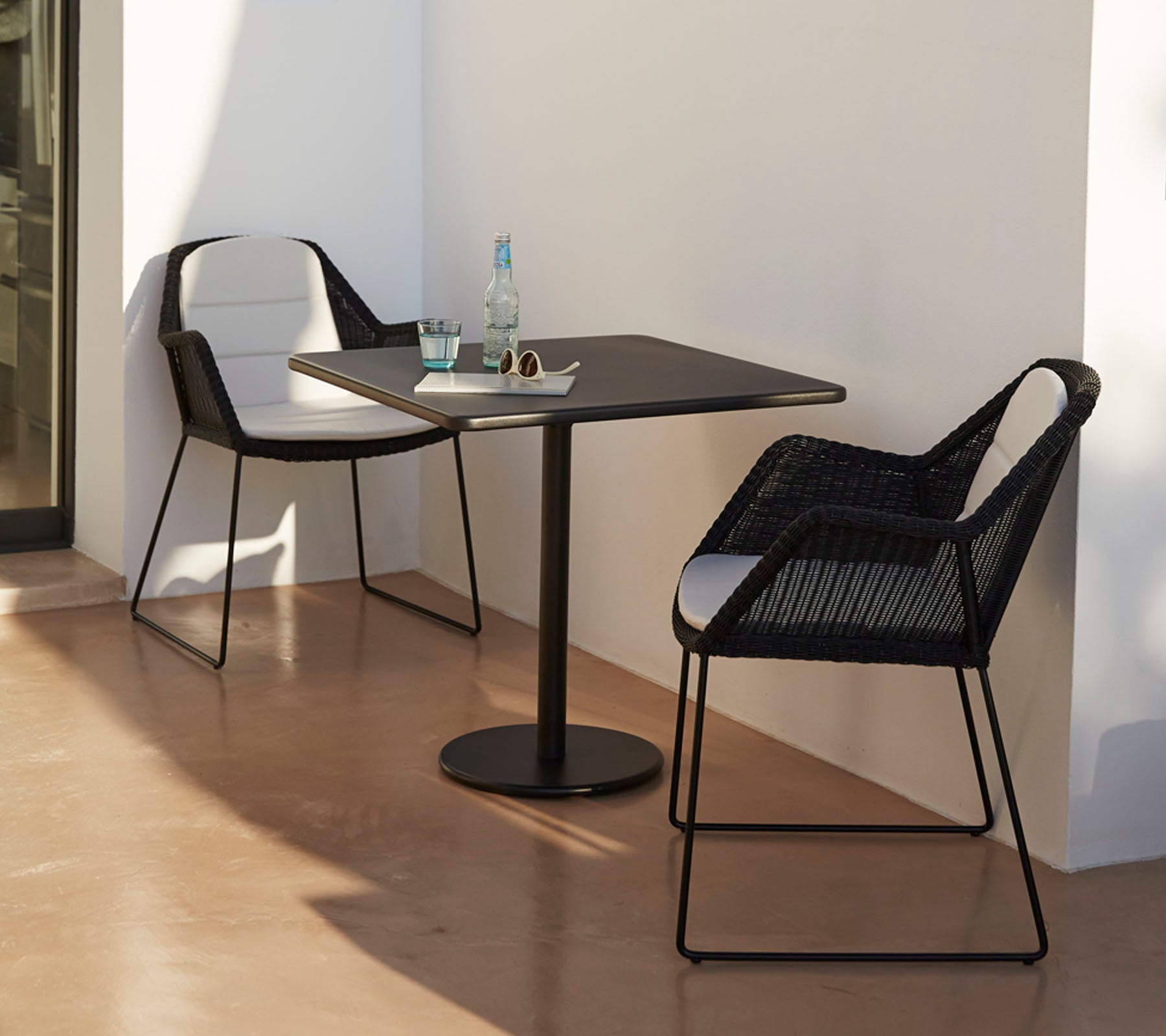 Moderne bord med to stilige svarte stoler, akkompagnert av en vannflaske og solbriller, satt mot en minimalistisk bakgrunn.
