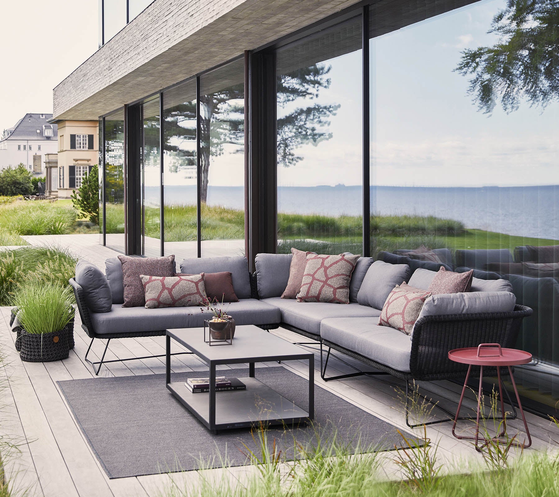 Moderne utendørs sitteområde med en seksjonert sofa med puter, et kaffebord og dekorative elementer blant frodig grønt.
