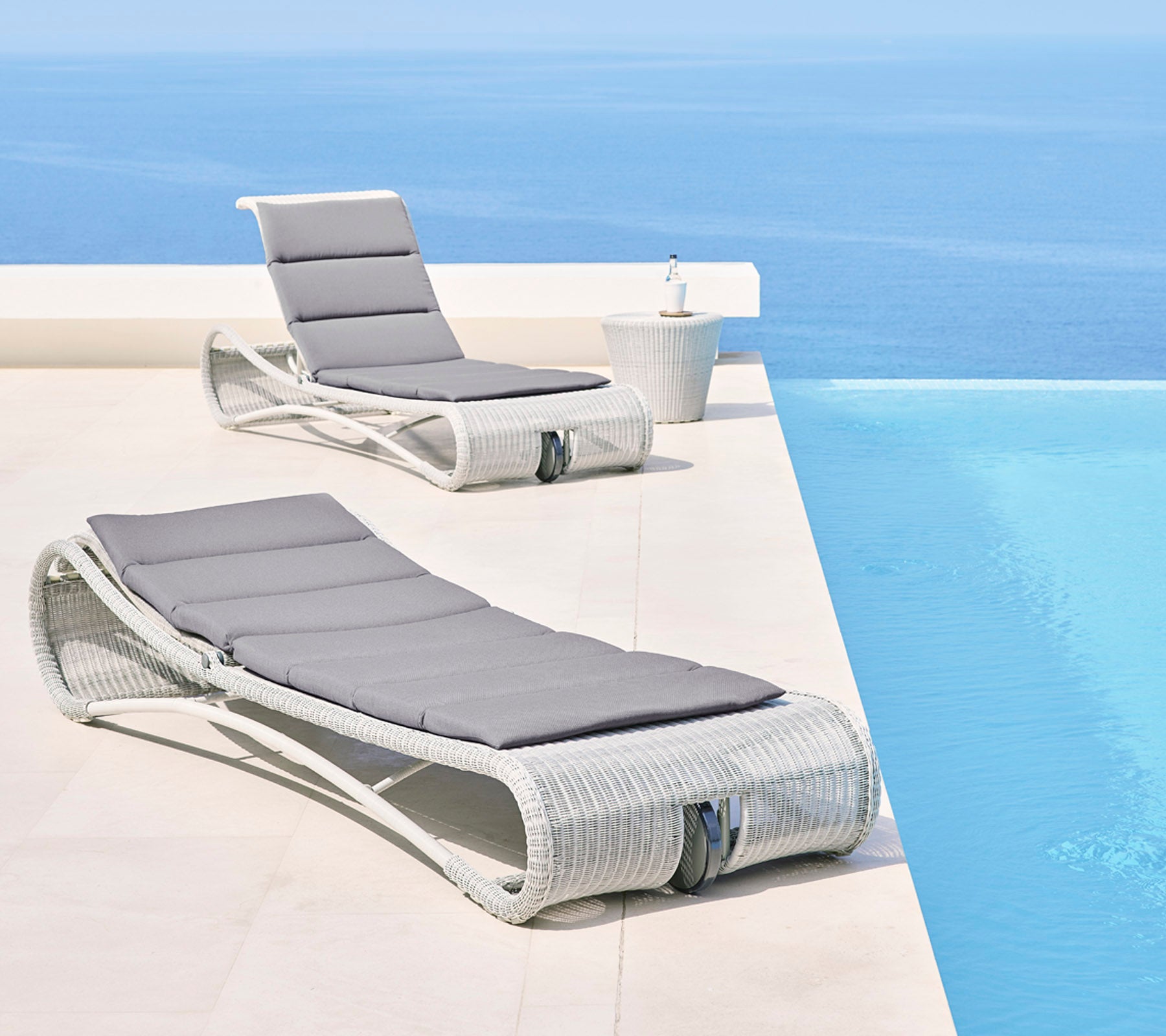 To stilige lenestoler med grå puter ved et rolig blått infinity-pool, som skaper et avslappende utendørsområde.