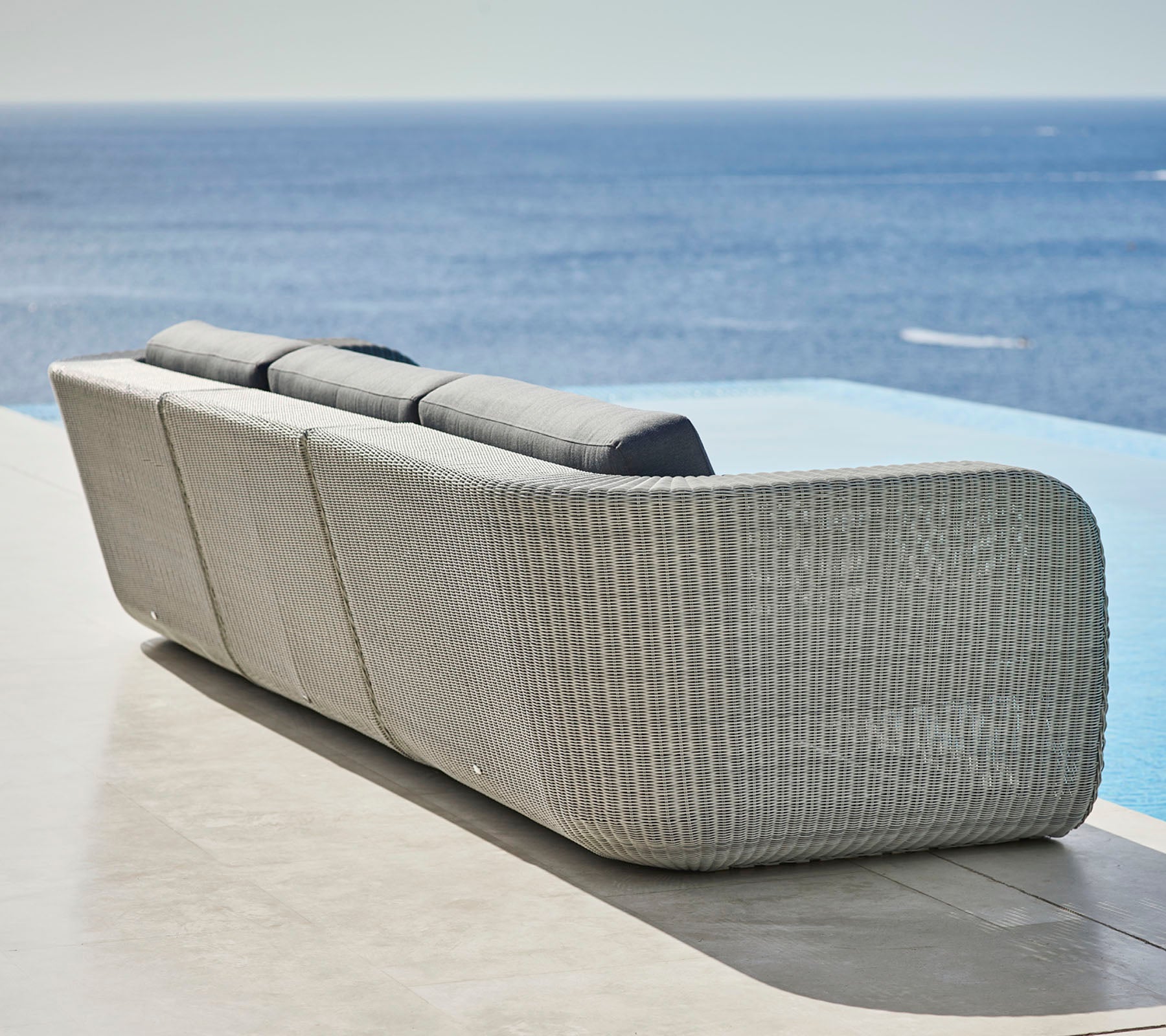 Stilig vevd sofa med buet design og myke puter, med utsikt over en rolig havutsikt. Ideell for avslapning og utendørs bruk.