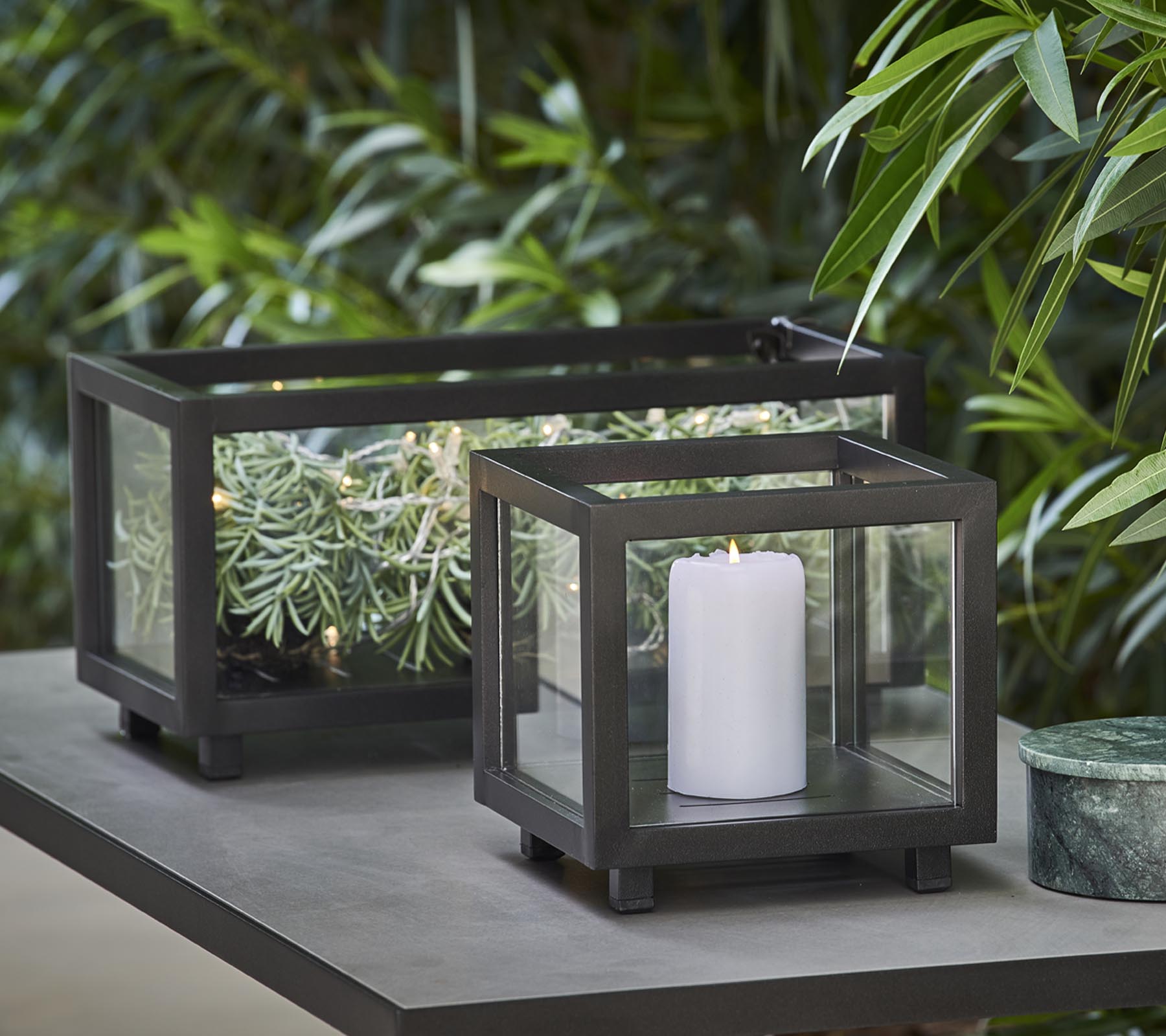 To stilige lykter med glassplater, den ene holder et hvitt stearinlys og den andre har grønne planter, plassert på et bord midt i frodig løvverk.