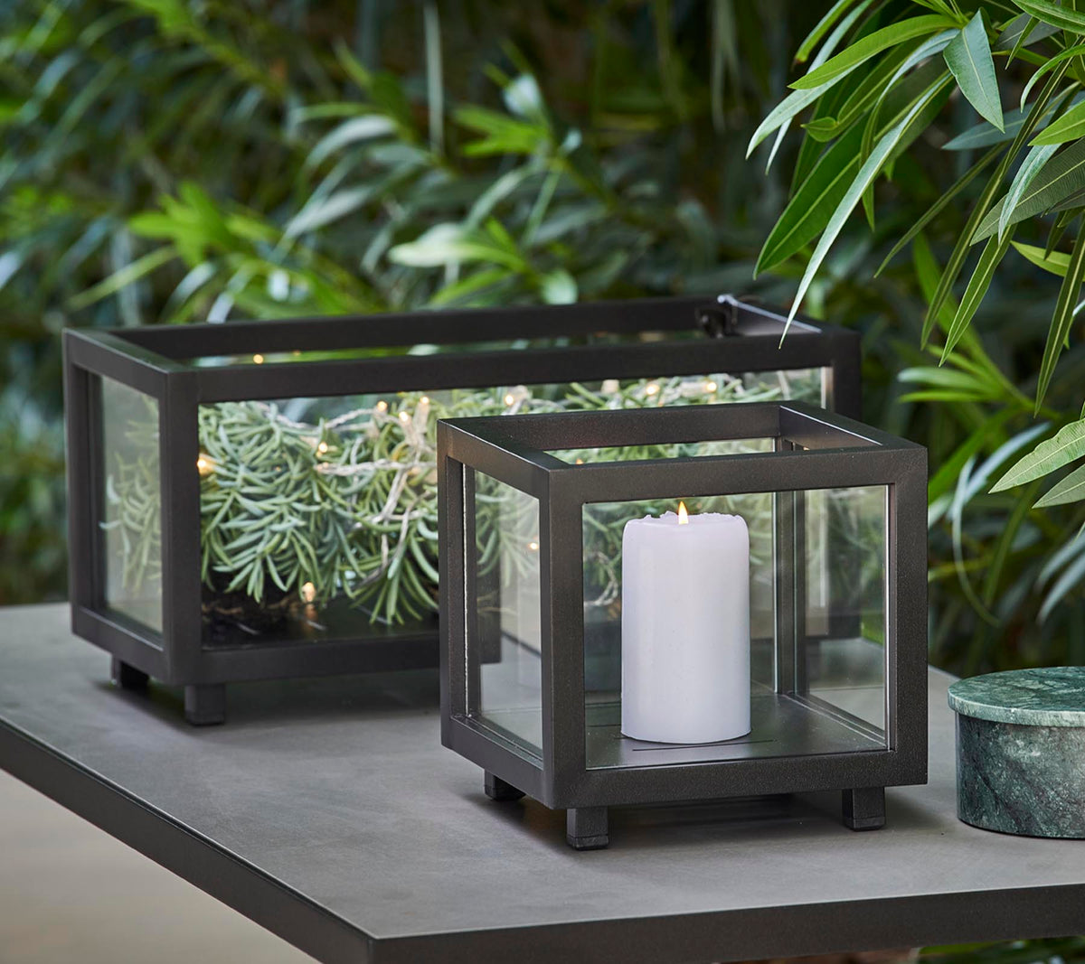 To stilige lykter med glassplater, den ene holder et hvitt stearinlys og den andre har grønne planter, plassert på et bord midt i frodig løvverk.
