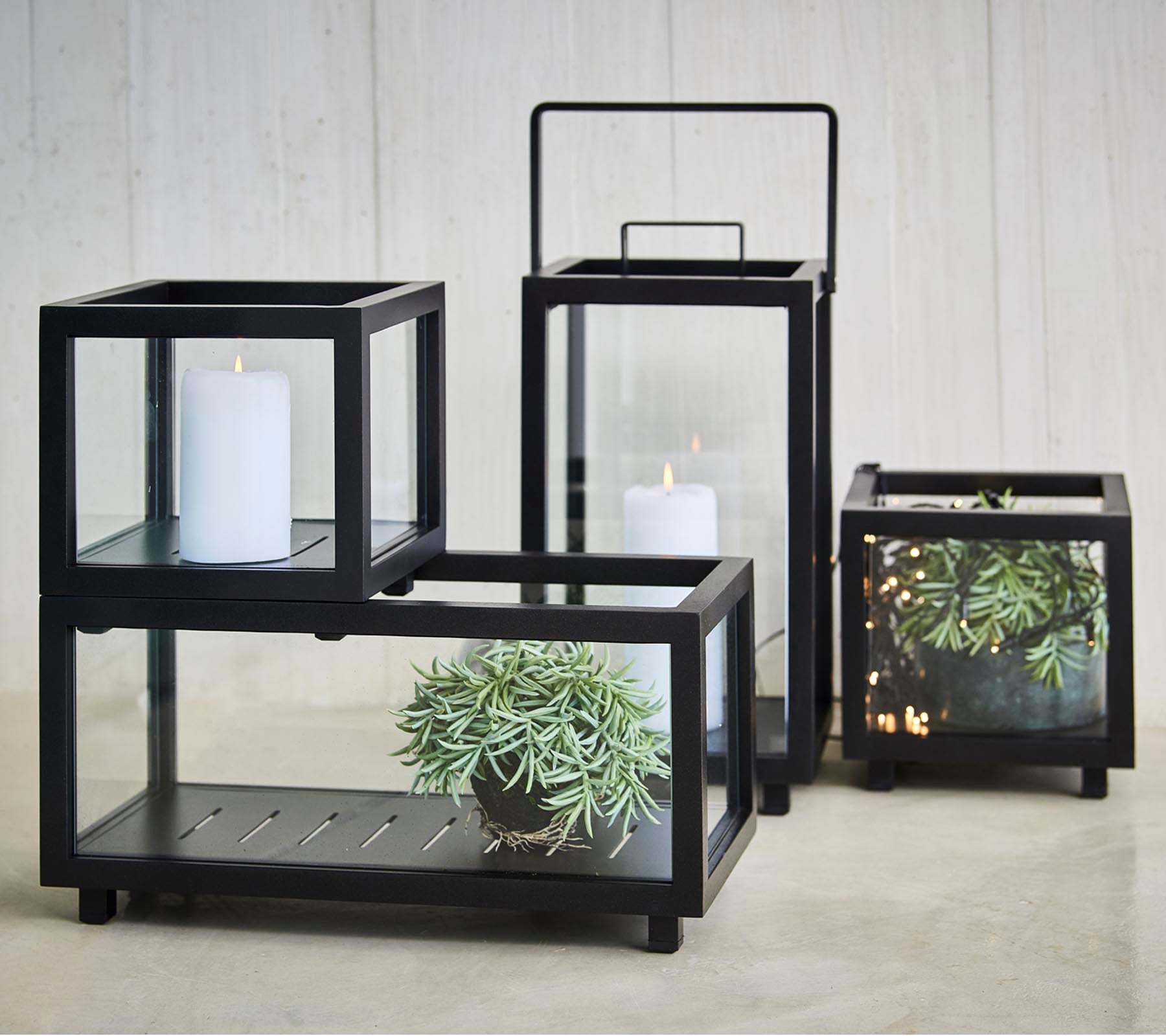 Et sett med elegante lysestaker med svarte rammer og glasspaneler som viser planter og stearinlys i forskjellige størrelser.