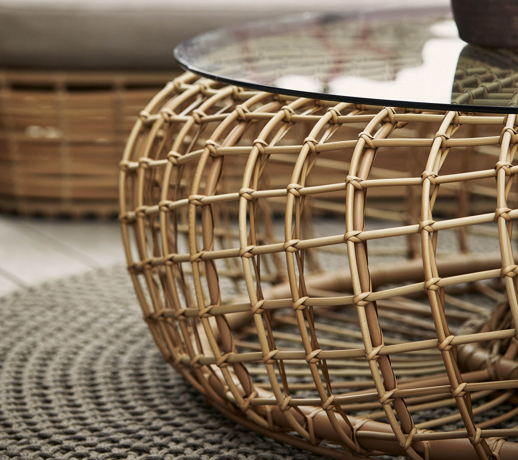 Rund kaffebord med en vevd rattanbase og glasskive, supplert med et teksturert rundt teppe.