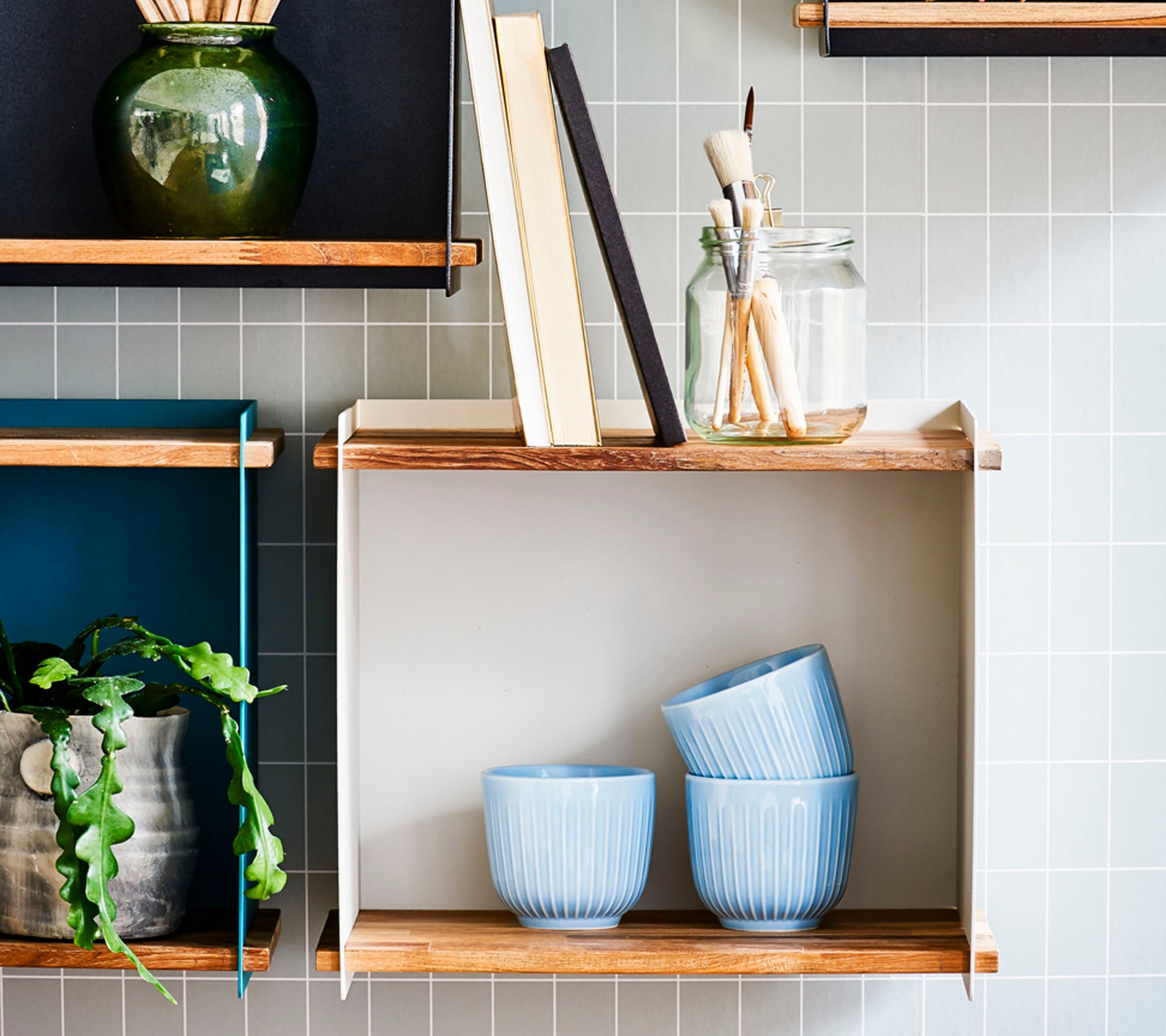 Hyller med dekorative gjenstander, inkludert blå skåler, bøker, et glass med pensler og en potteplante mot en flislagt bakgrunn.