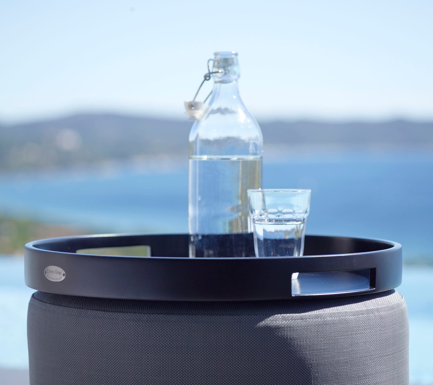En svart rund bakke holder et glass vann og en glassflaske, satt mot en naturskjønn bakgrunn av havet og fjerne åser.