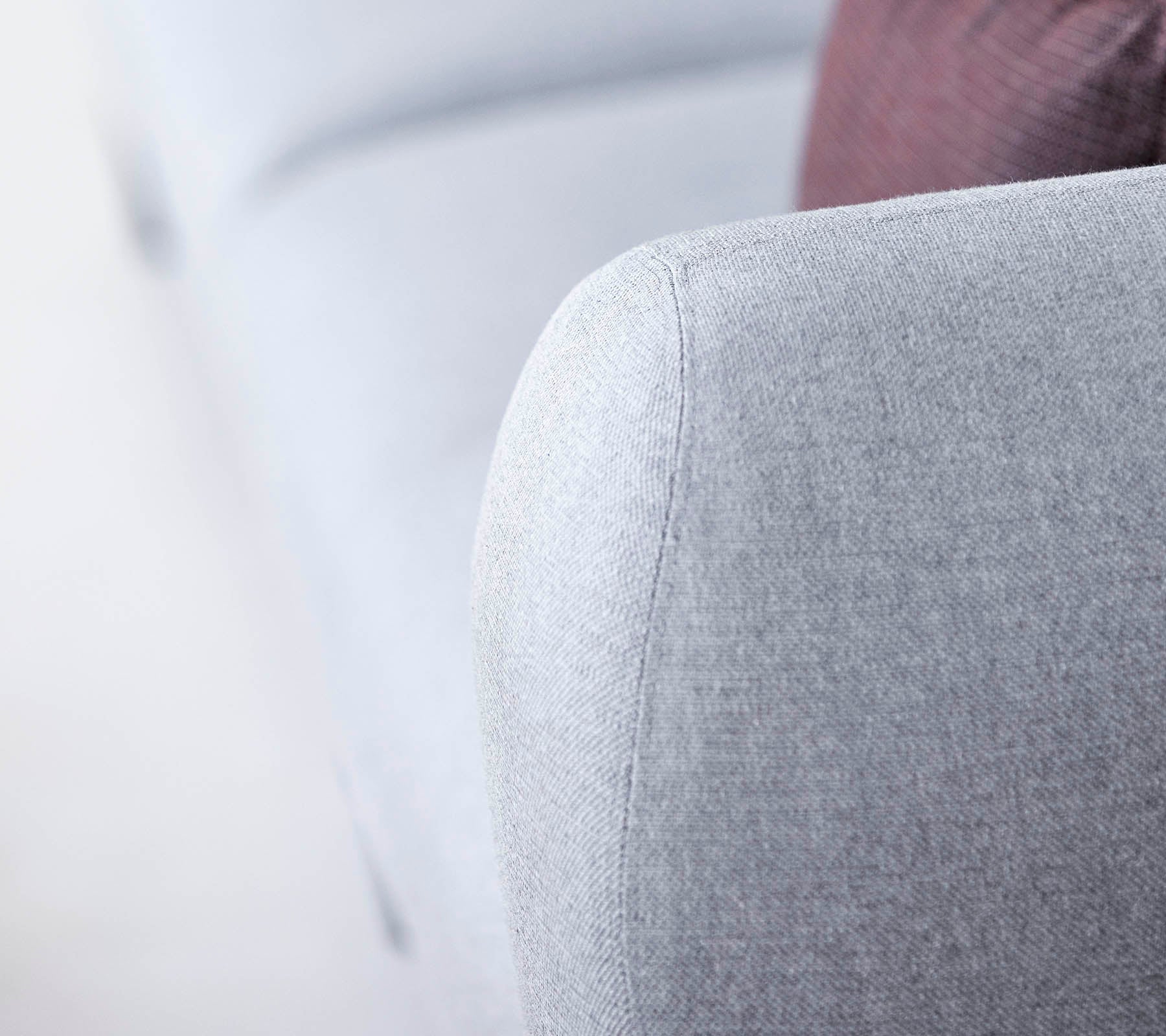 Nærbilde av et lysegrå sofaarm, med et teksturert stoff og et mykt, avrundet design, med et hint av en mørkere pute i bakgrunnen.