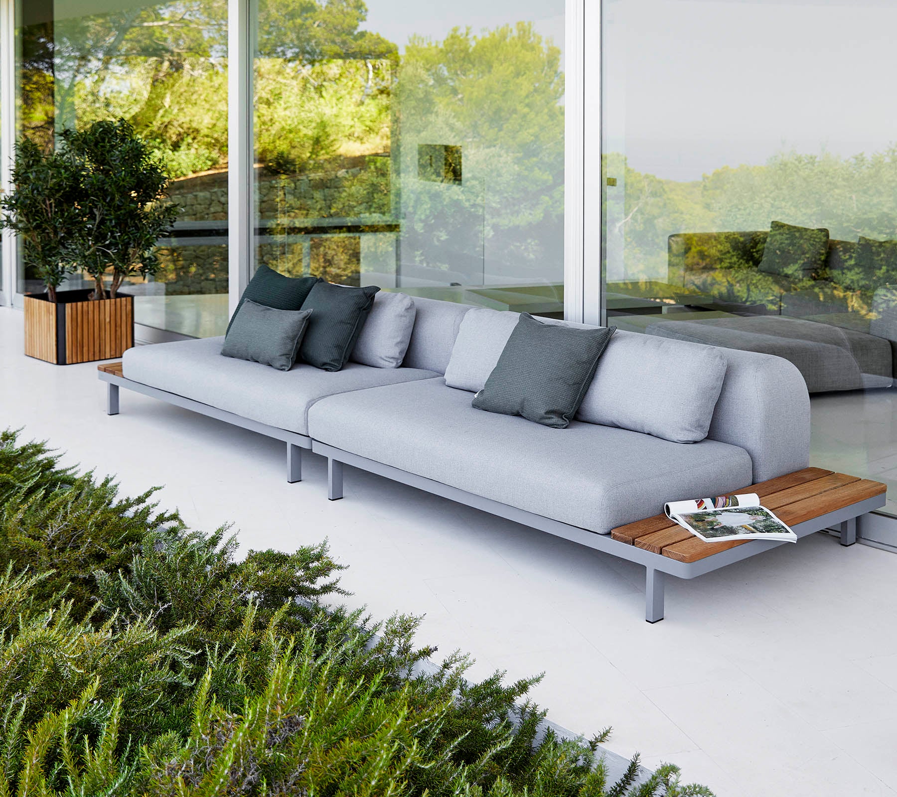 Moderne sofa med lysegrå puter og tredekor, akkompagnert av dekorative puter og omgitt av grønt.